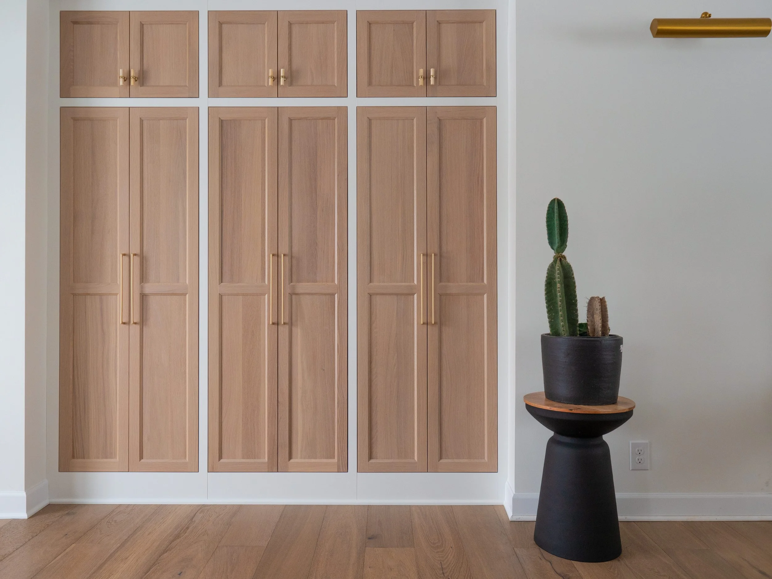 A wooden wardrobe with six doors and gold handles, against a white wall. To the right, a black planter with a tall cactus with three main stems, placed on a black and wood side table. The floor is wooden.