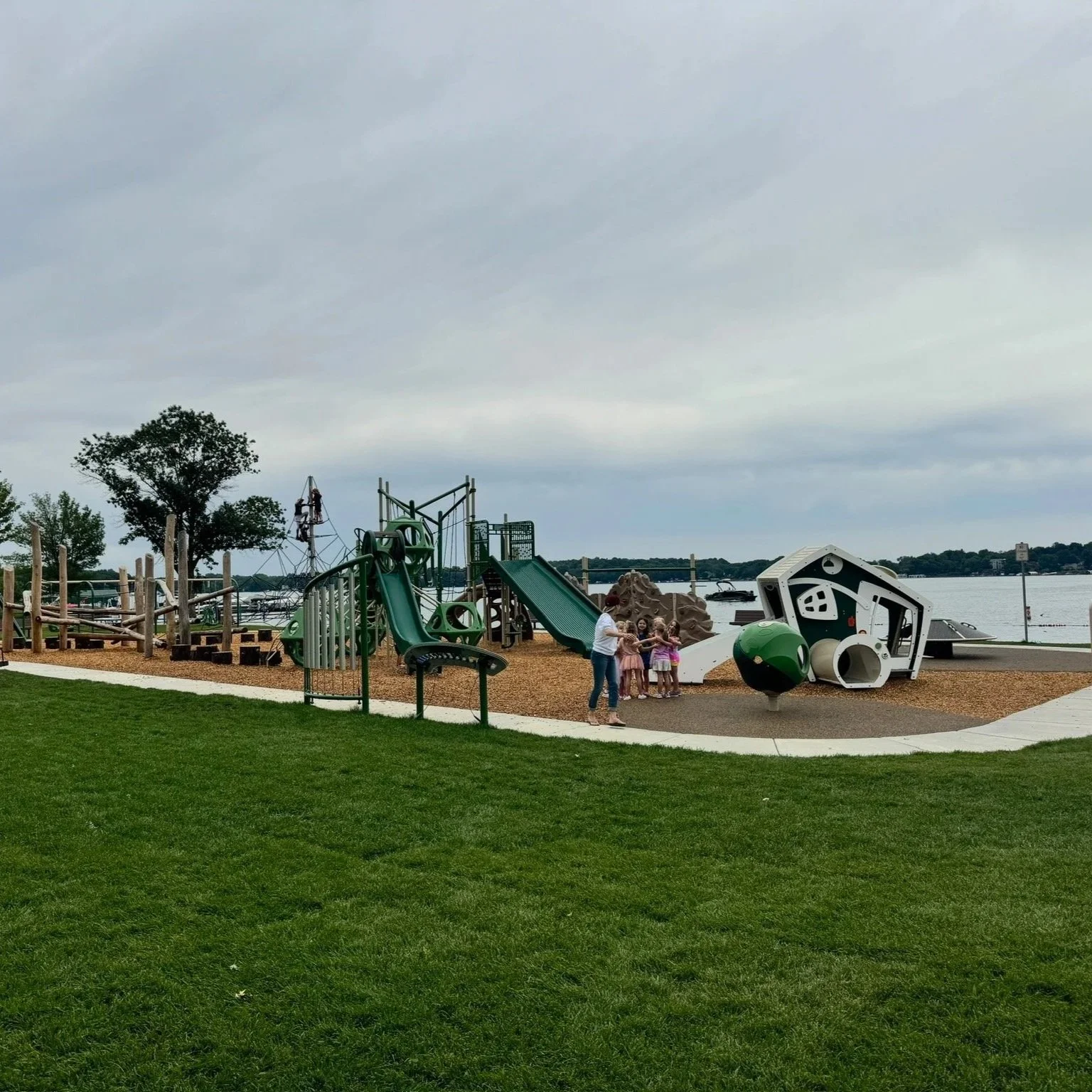 Playground near a waterfront with a green slide, climbing structures, a large space-themed art installation, and children playing with an adult.