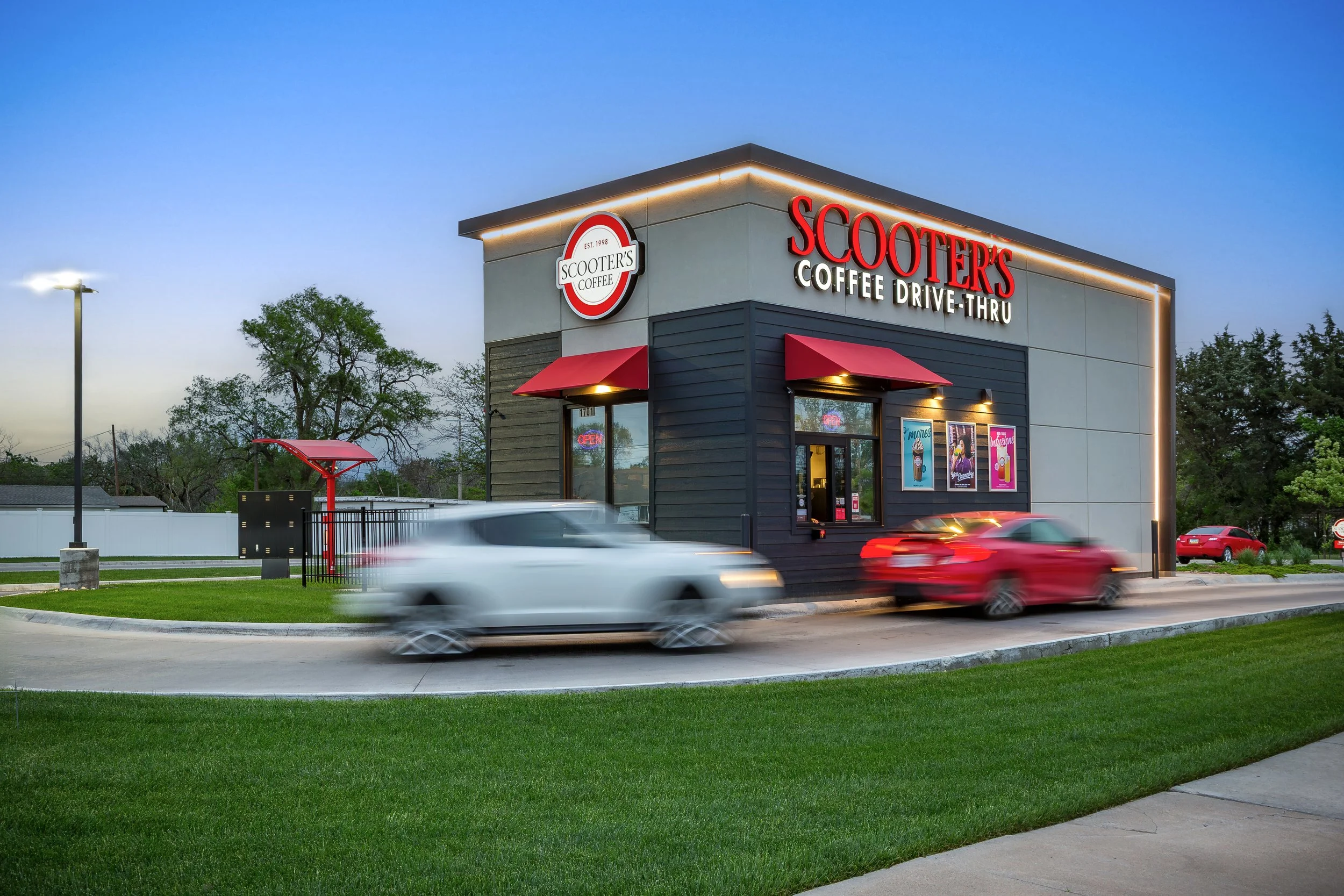 Professional photography shoot of A fast-food restaurant called Scooter's Coffee with a drive-thru, showing cars in motion during the evening with a clear sky and trees in the background.