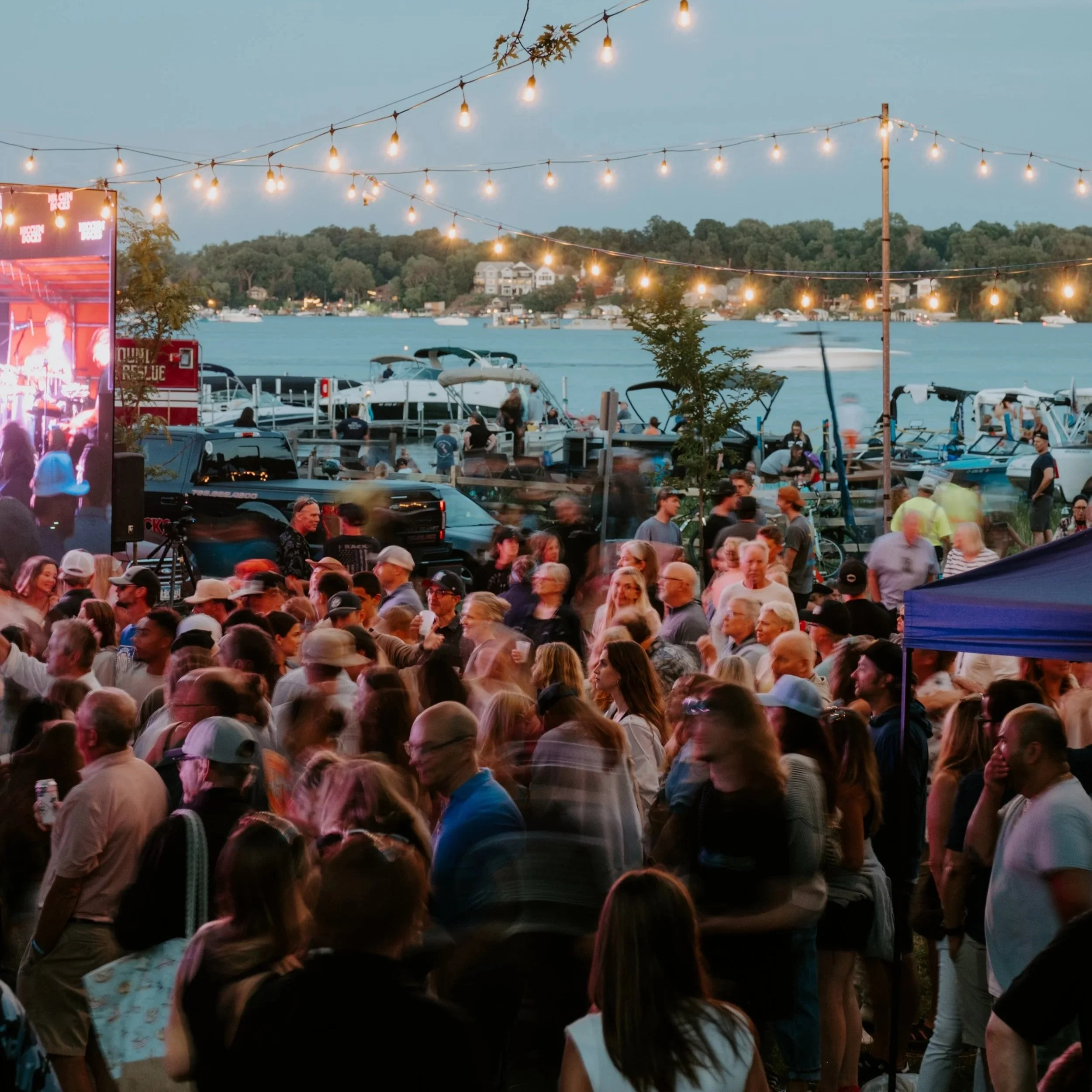 Crowd gathered at an outdoor event near a marina with boats, water, and string lights overhead, during early evening.