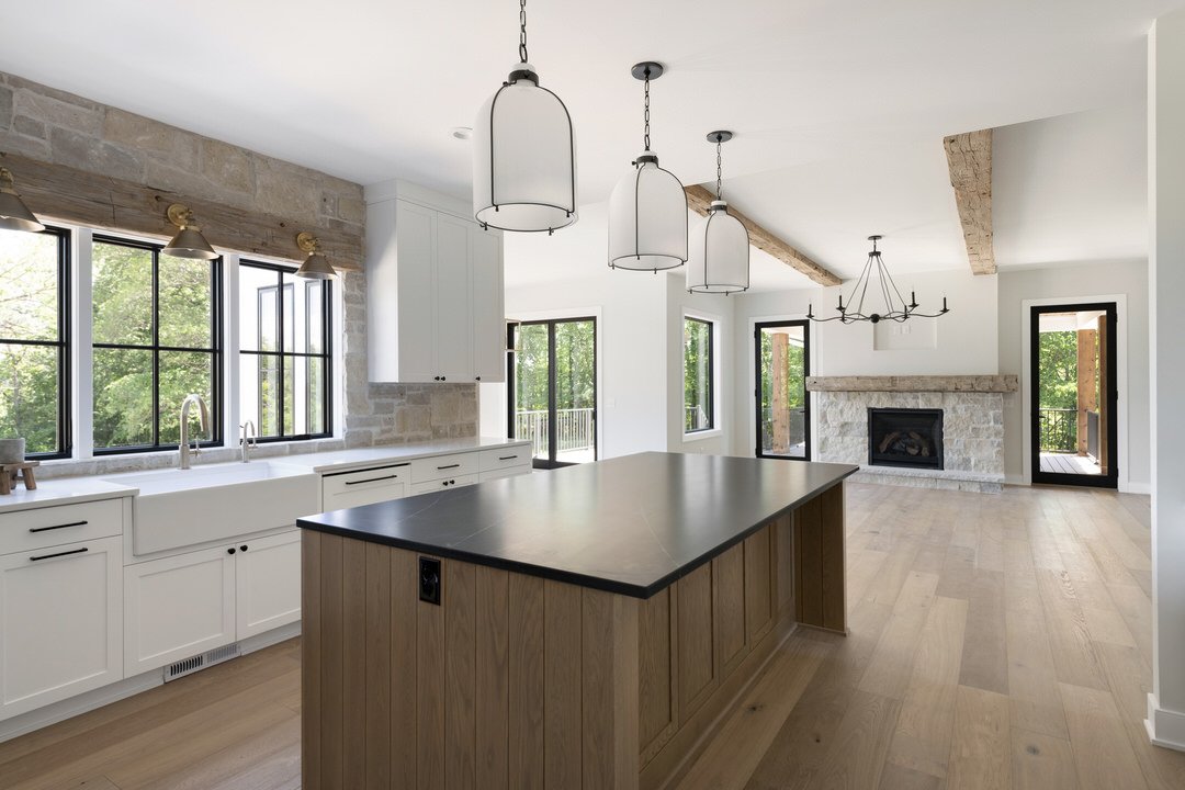 Modern kitchen with white cabinets, black window frames, a large island with a dark countertop, hardwood floors, and a living room area with a stone fireplace and large windows.