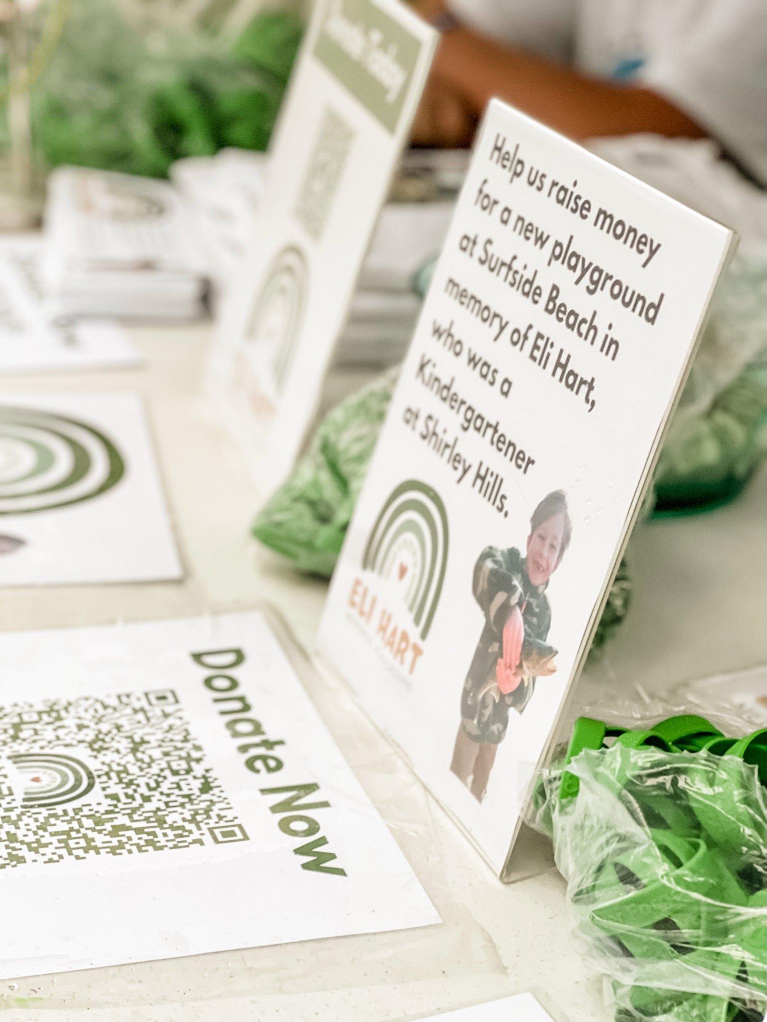 A sign on a table reads: "Help us raise money for a new playground at Surfside Beach in memory of Eli Hart, who was a kindergartner at Shirley Hills." Behind the sign, there is a photo of a young girl. There are also some QR codes and green plastic wristbands on the table.