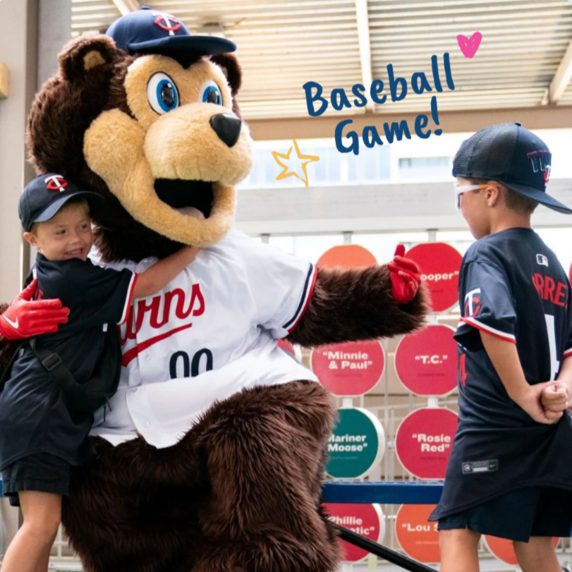 Children with a mascot in a baseball stadium, smiling and posing for a photo, with a sign that says 'Baseball Game!'