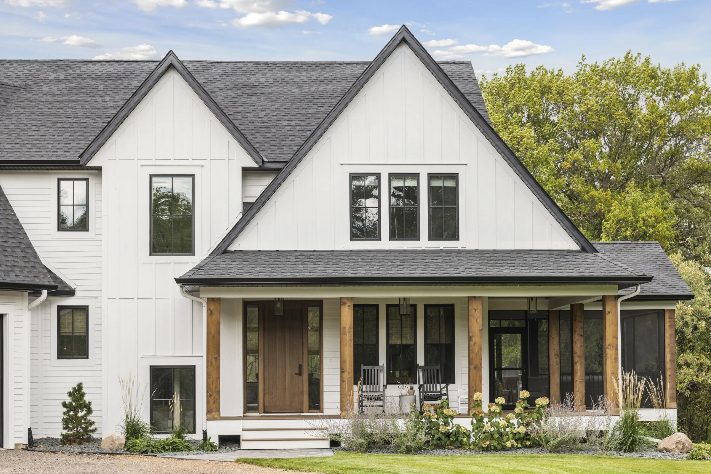Modern two-story house with white siding, black window frames, a front porch with wooden columns, and a landscaped yard with greenery and rocks.