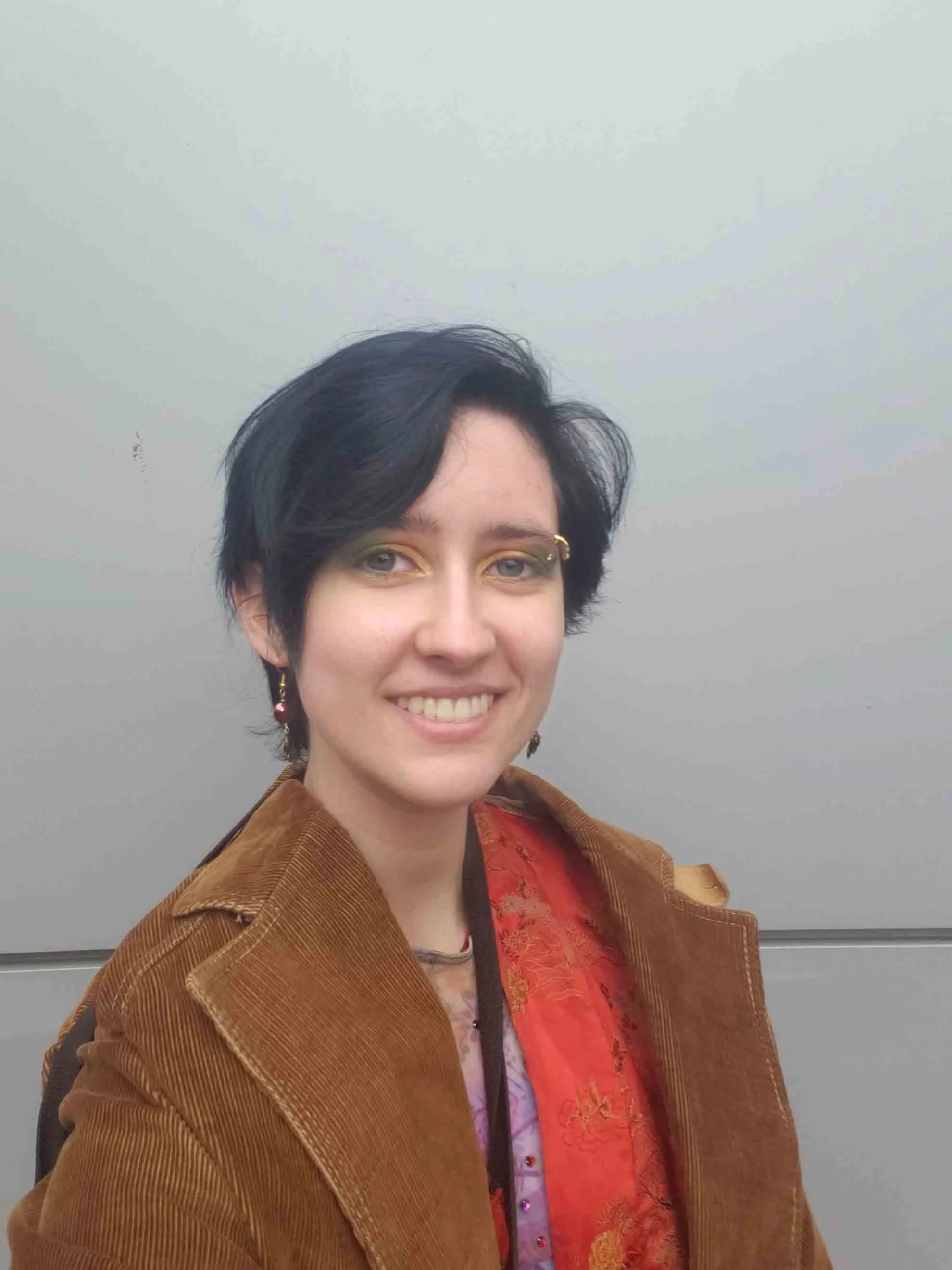Blue-eyed dark haired white trans man smiling, wearing a brown coat and colorful eyeshadow.