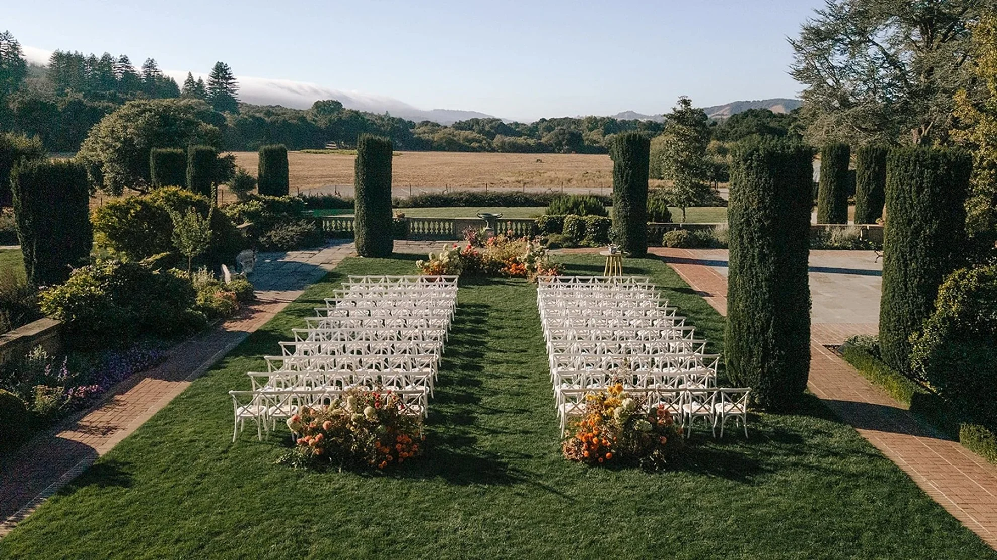 Formal garden wedding ceremony featuring statement greenery and floral installations, elegant white chairs lining the aisle, and a lush landscaped backdrop, thoughtfully designed by a luxury full-service wedding planning team.