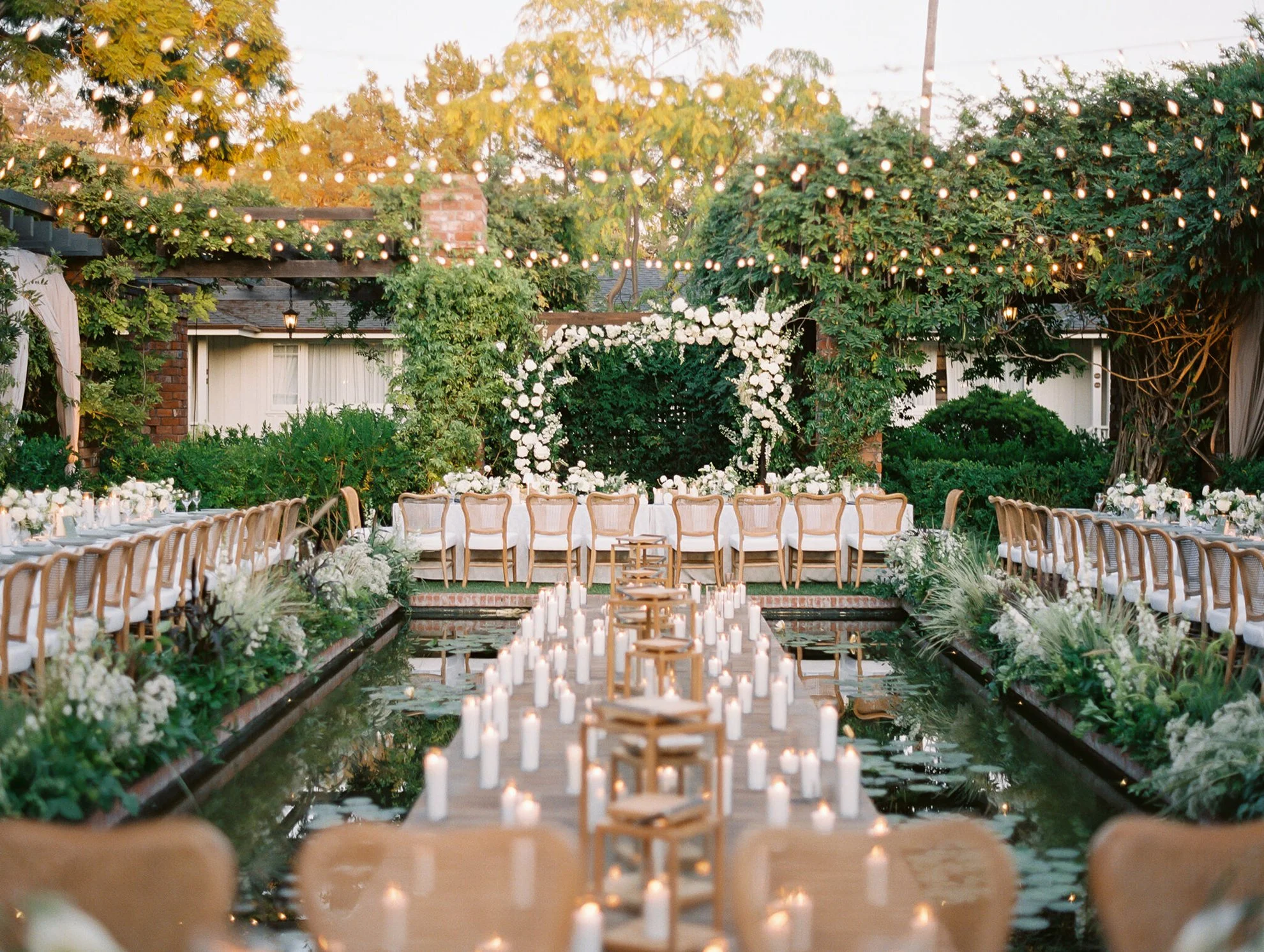 Elegant outdoor garden wedding reception featuring candlelit dining tables, rattan chairs, lush greenery, and green and white floral arrangements set beside a serene pond, designed by a luxury full-service wedding planner.