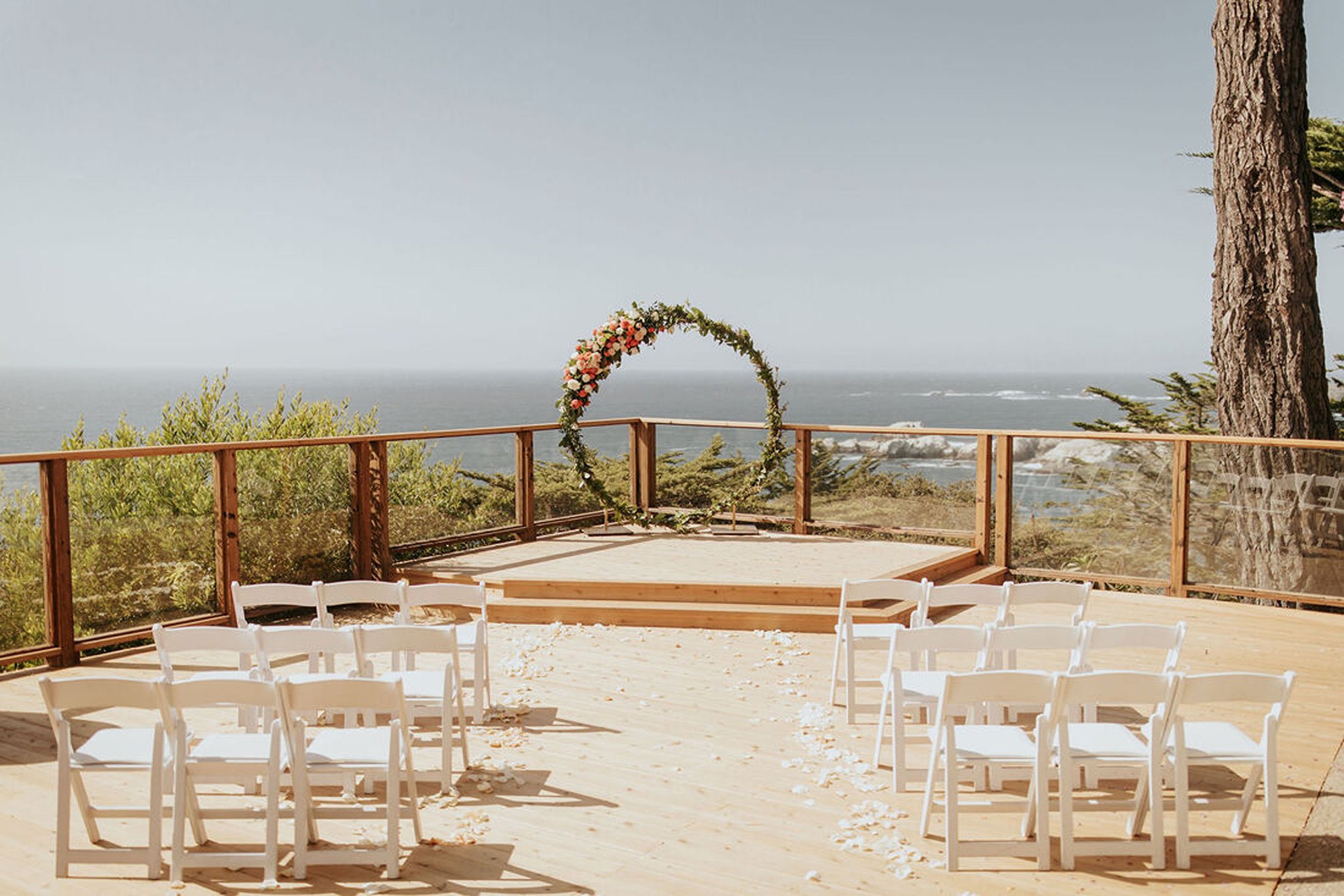 Minimalist outdoor coastal wedding ceremony featuring a circular floral arch, white guest chairs lining the aisle, and sweeping scenic ocean views, thoughtfully designed by a full-service luxury wedding planning team.