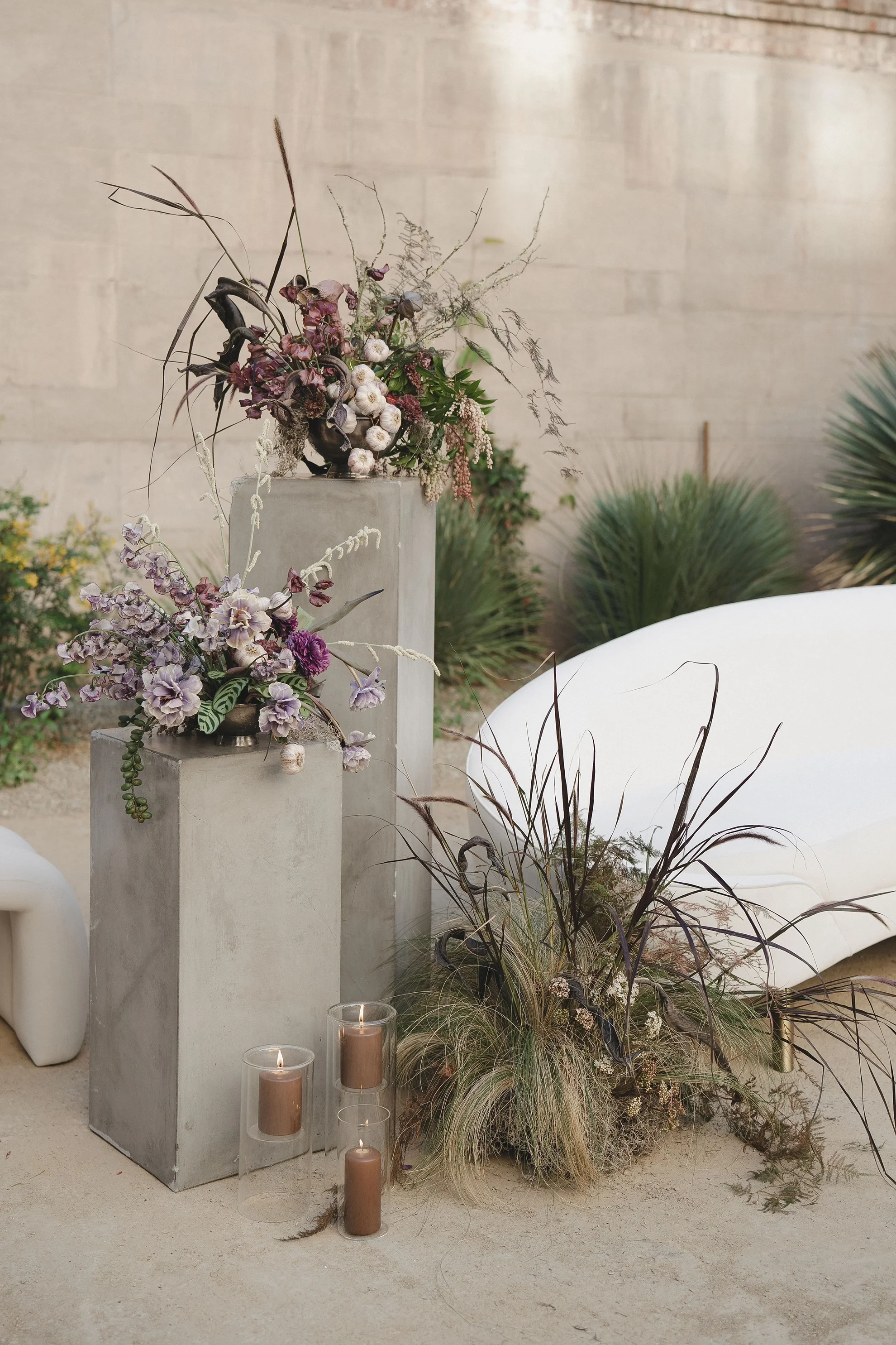 Purple and cream modern florals with a garlic centerpiece on a concrete pedestal, creating a playful and unexpected wedding design moment with layered color and texture