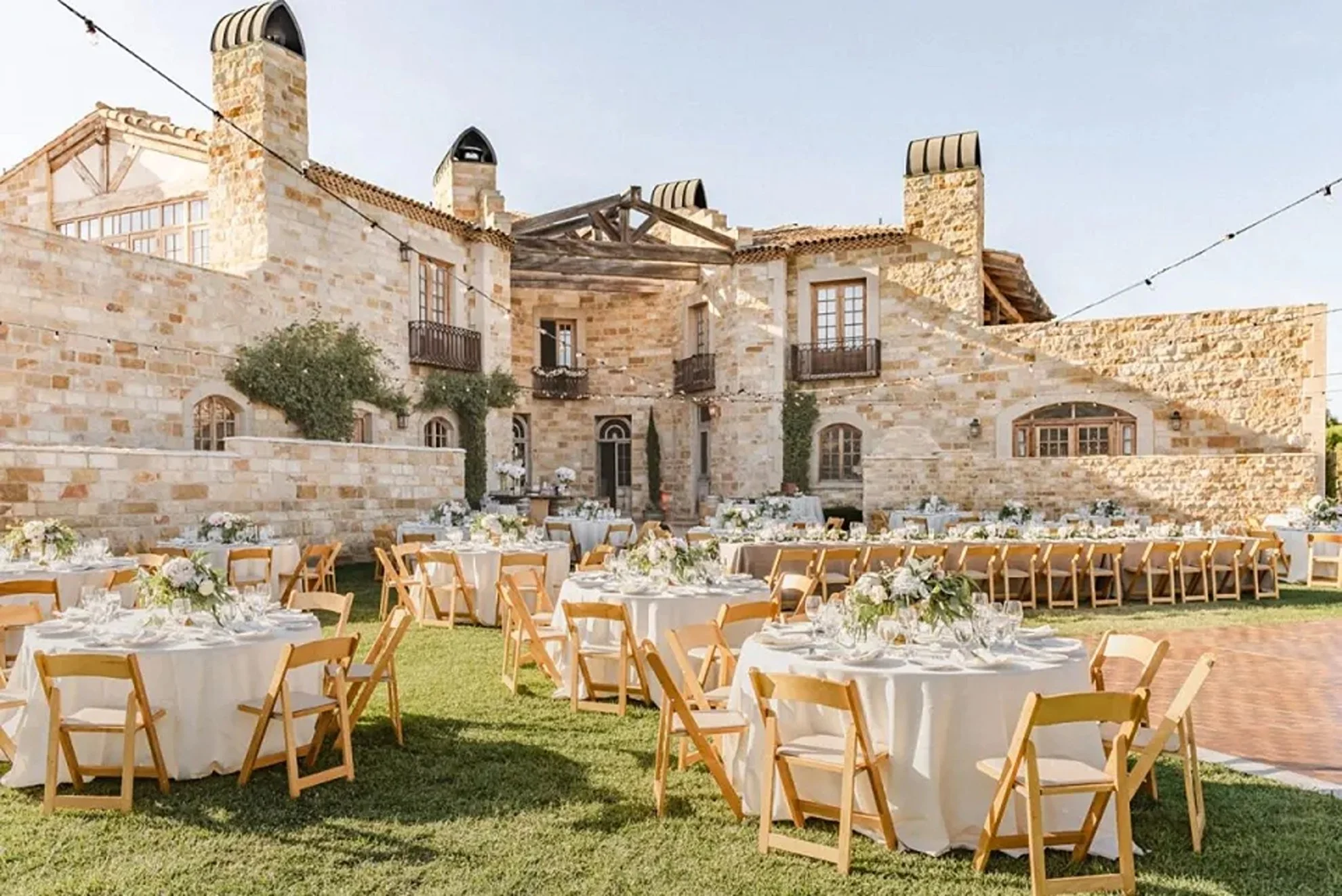 Outdoor wedding reception set against a European-inspired architectural estate, featuring wooden dining chairs and elegant floral centerpieces, thoughtfully designed and produced by a luxury full-service wedding planning team.