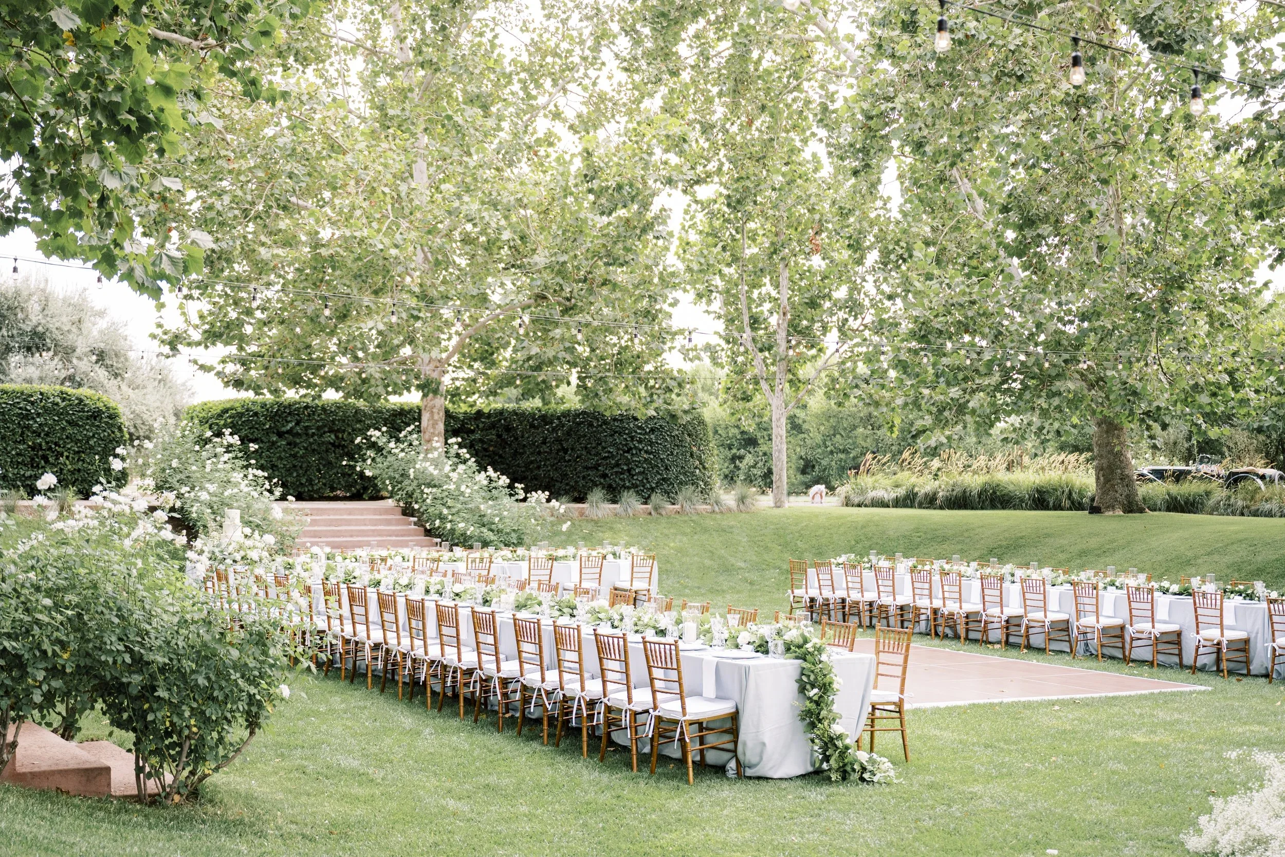 Elegant outdoor wedding reception set on lush green grass, featuring abundant greenery, wooden dining chairs, and a flowing green and white floral garland, thoughtfully designed by a luxury full-service wedding planning team.