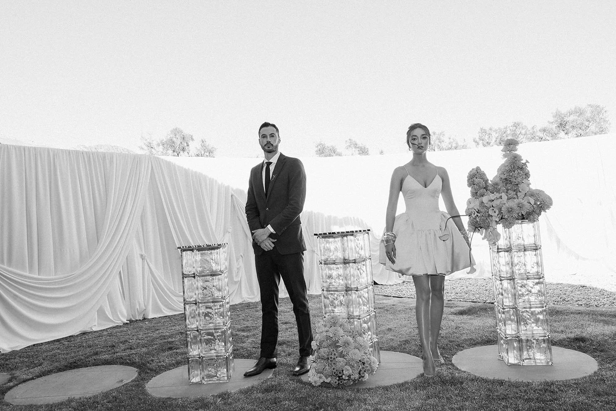 Bride in a Danielle Frankel mini dress standing in front of a draped wall, showcasing a fashion-forward and modern wedding look with bold, intentional styling