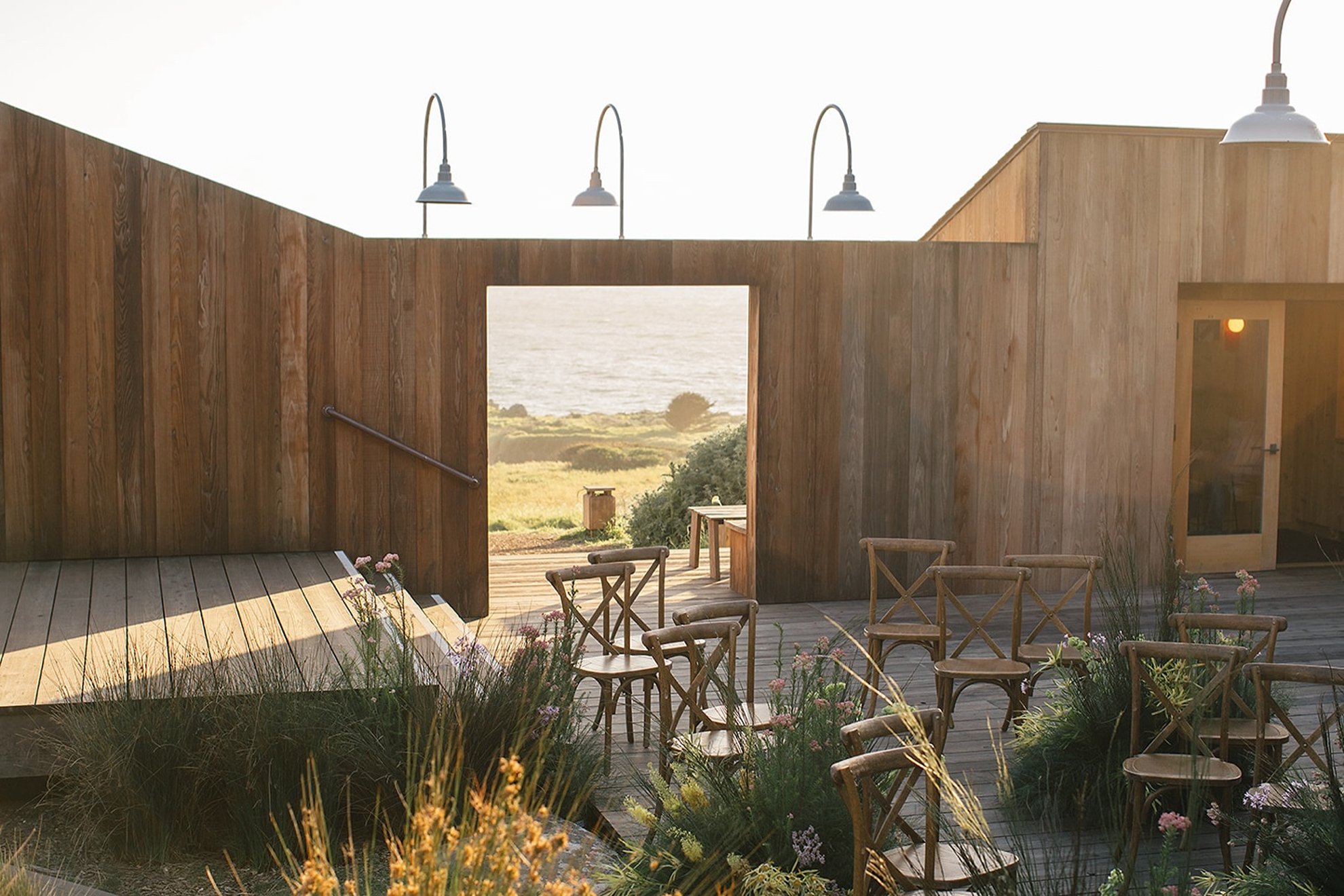 Modern coastal lodge wedding reception set within a landscaped outdoor setting, featuring lush floral arrangements and wooden dining chairs, thoughtfully designed and produced by a luxury full-service wedding planning team.