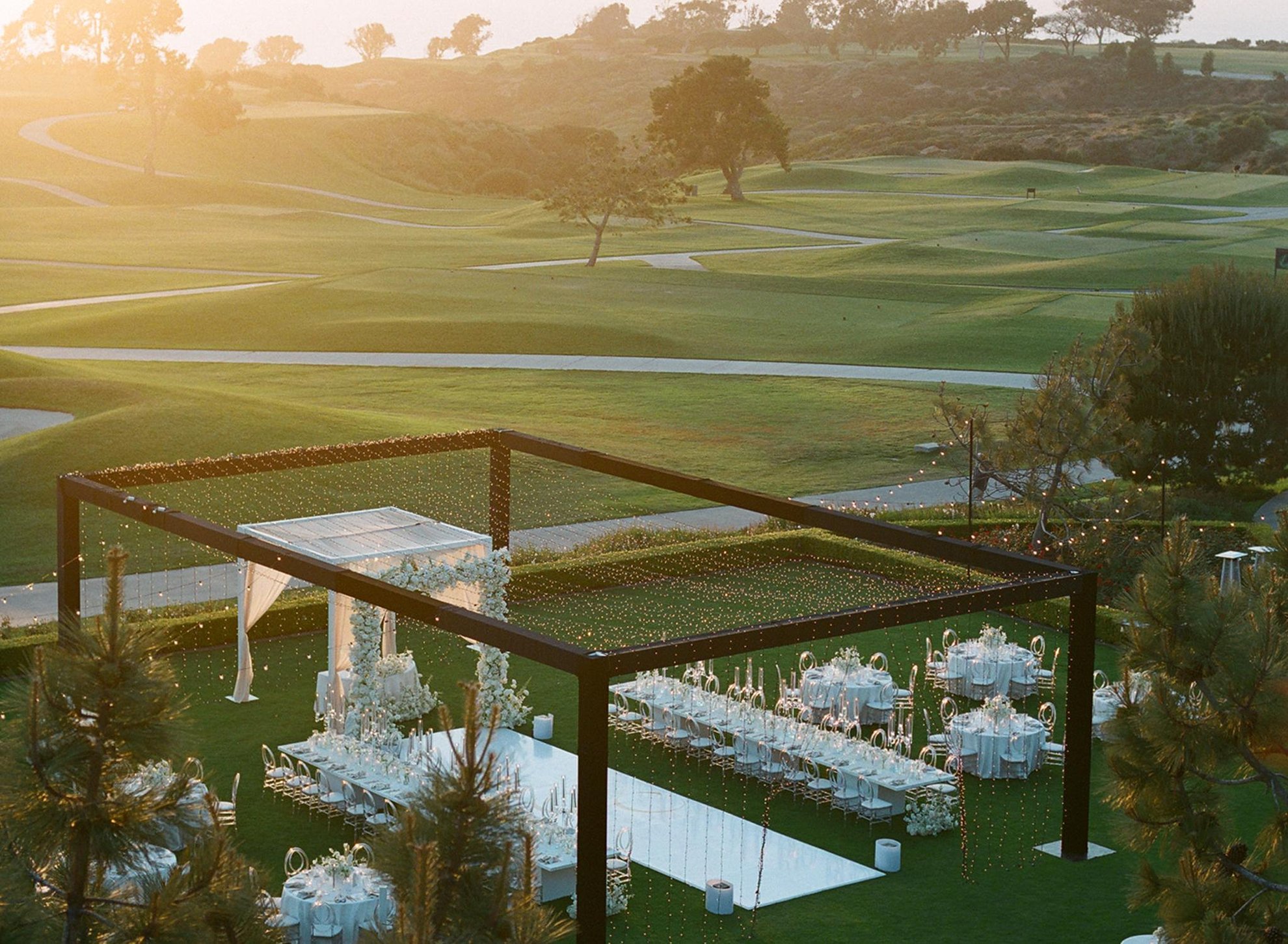 Classic outdoor wedding reception overlooking a manicured golf course, featuring a custom string light structure, clean modern tablescapes, and elegant white floral arrangements, thoughtfully designed by a luxury full-service wedding planning team.
