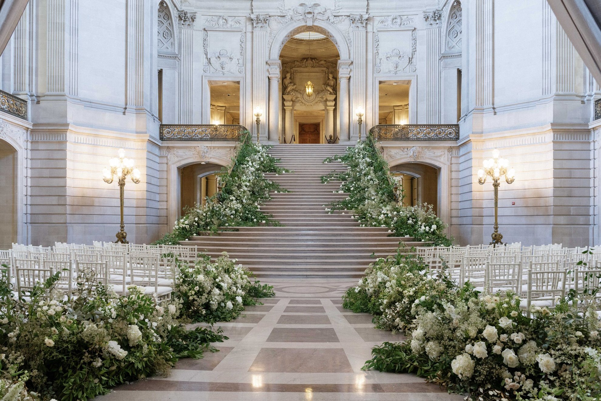 Elegant city hall wedding ceremony setup featuring grand elevated floral installations lining the aisle and staircase, set against striking architectural details, designed and produced by a luxury full-service wedding planning team.