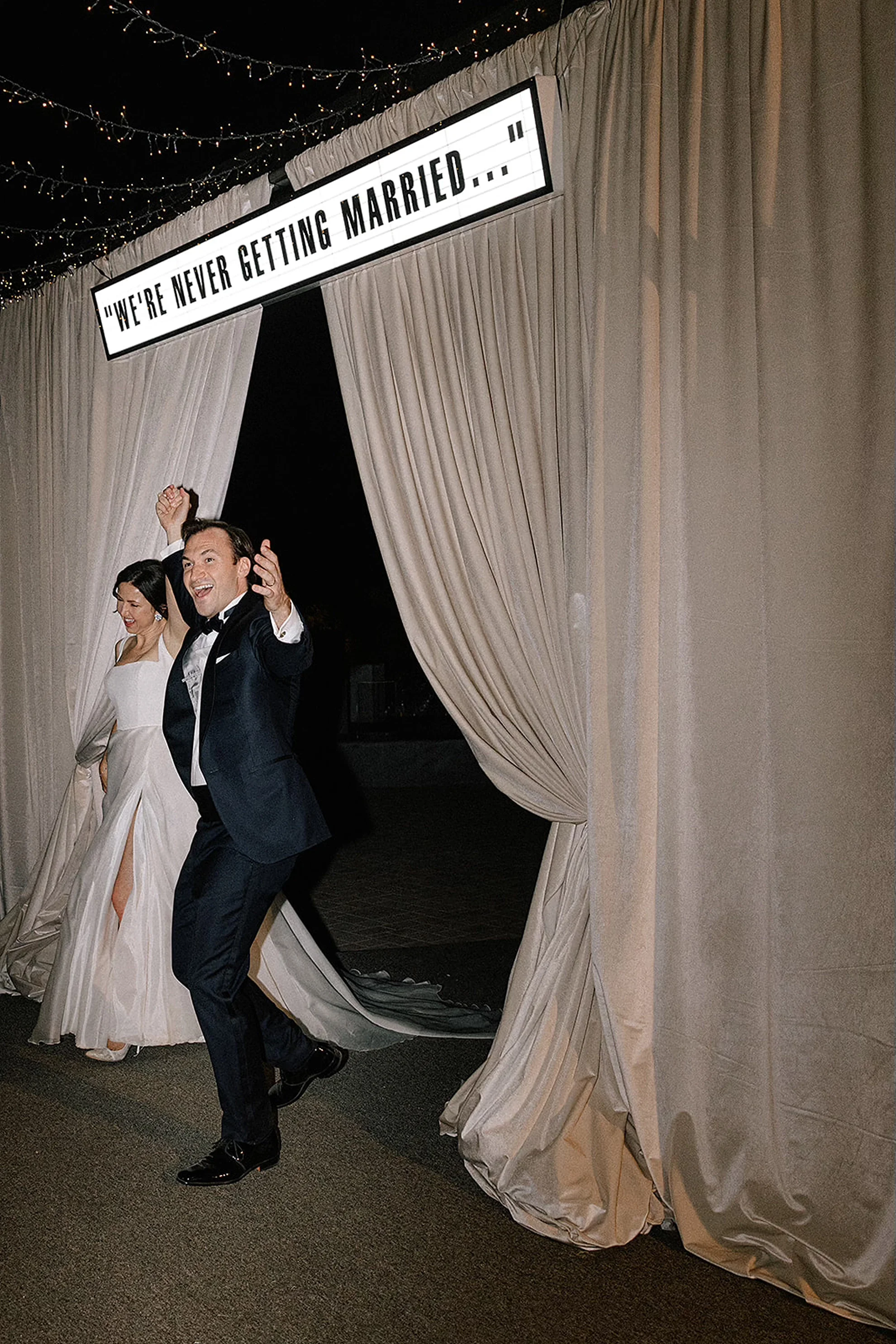 Joyful couple entering a tented wedding reception beneath a “we’re never getting married” sign, designed by Mandy Marie Events.