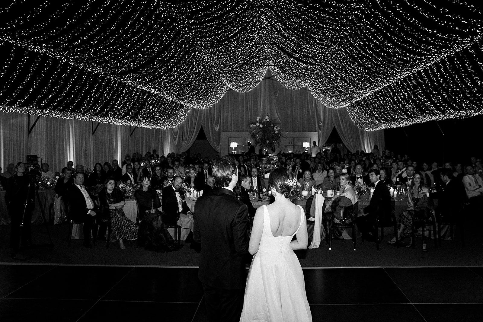 “Couple dancing on stage with friends and family to a live band beneath twinkle lights in an open-air wedding tent in Scottsdale, Arizona.”