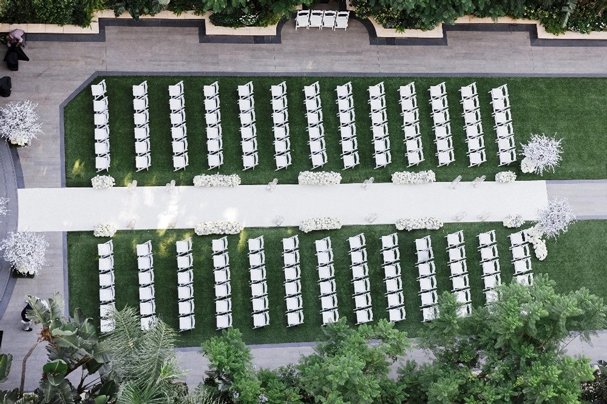 Grand outdoor wedding ceremony featuring white guest chairs, a long white aisle, elevated white floral installations, and lush green landscape surroundings, expertly designed and produced by a luxury full-service wedding planning team.