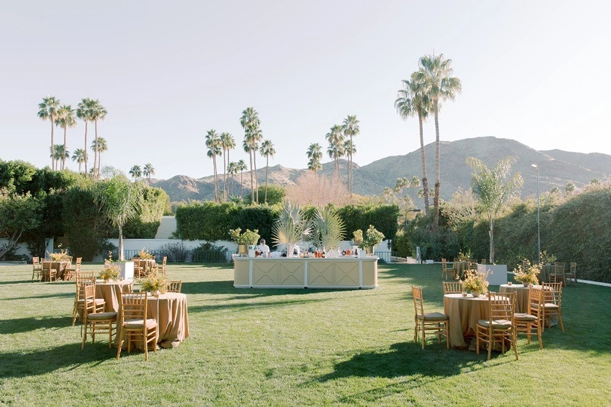 Outdoor wedding reception dinner set within a lush green landscape, featuring wooden dining chairs, a thoughtfully styled bar setup, and cohesive floral arrangements throughout, designed and produced by a luxury full-service wedding planning team.