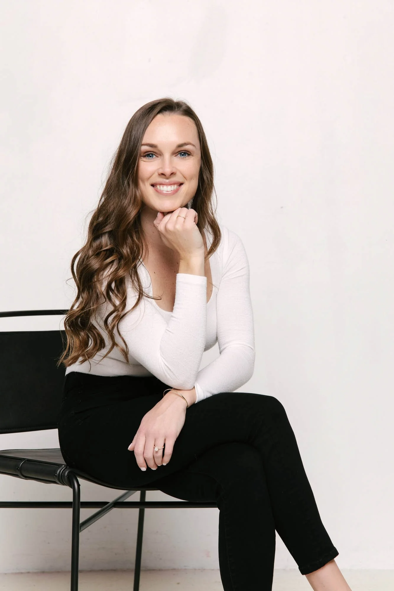 A woman with long, wavy brown hair, smiling, sitting on a black chair against a white background, resting her chin on her hand.