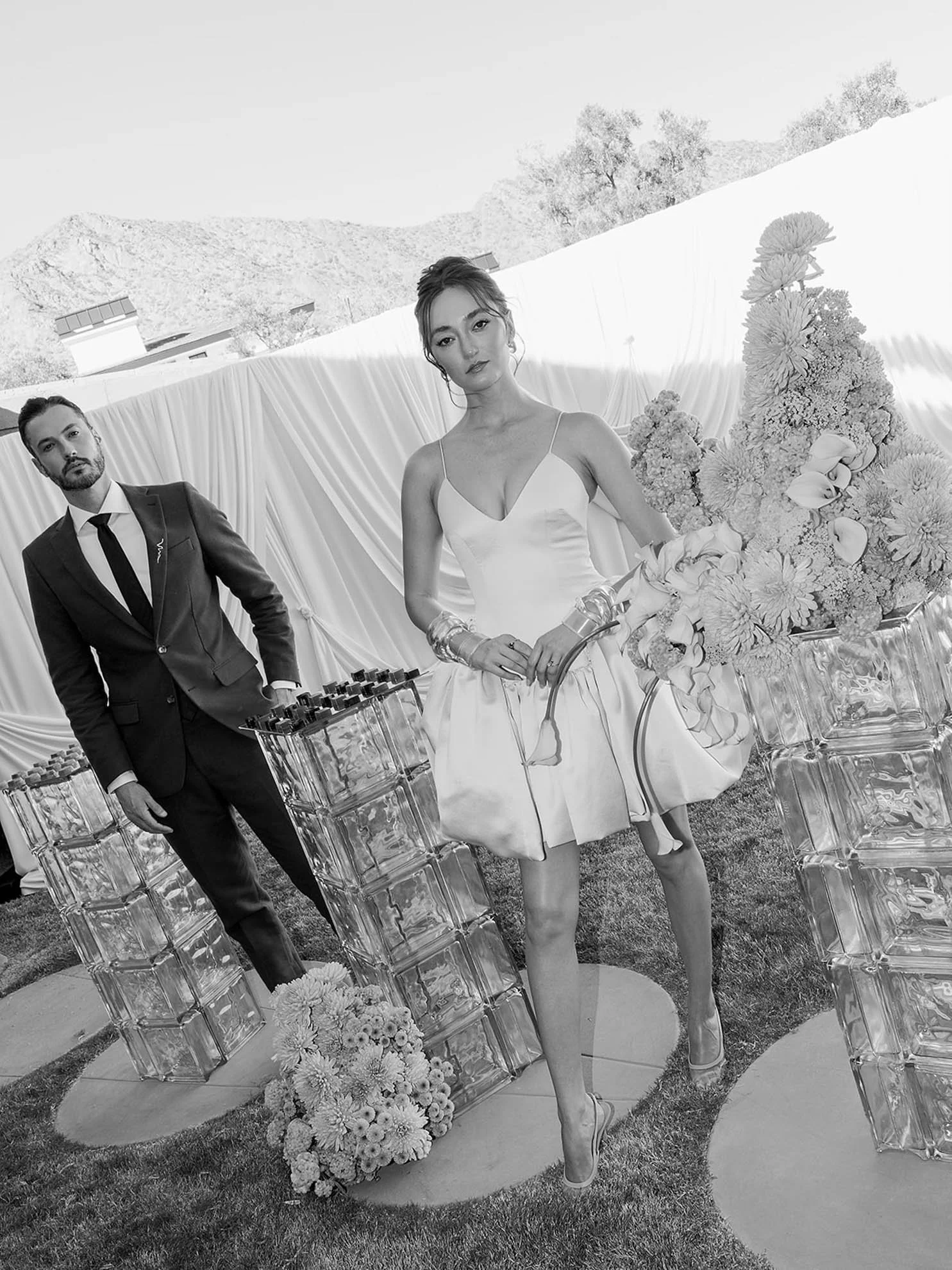 A black and white photo of a woman in a wedding dress standing in front of a floral arrangement at an outdoor wedding ceremony, with a man in a suit standing nearby and mountains visible in the background.