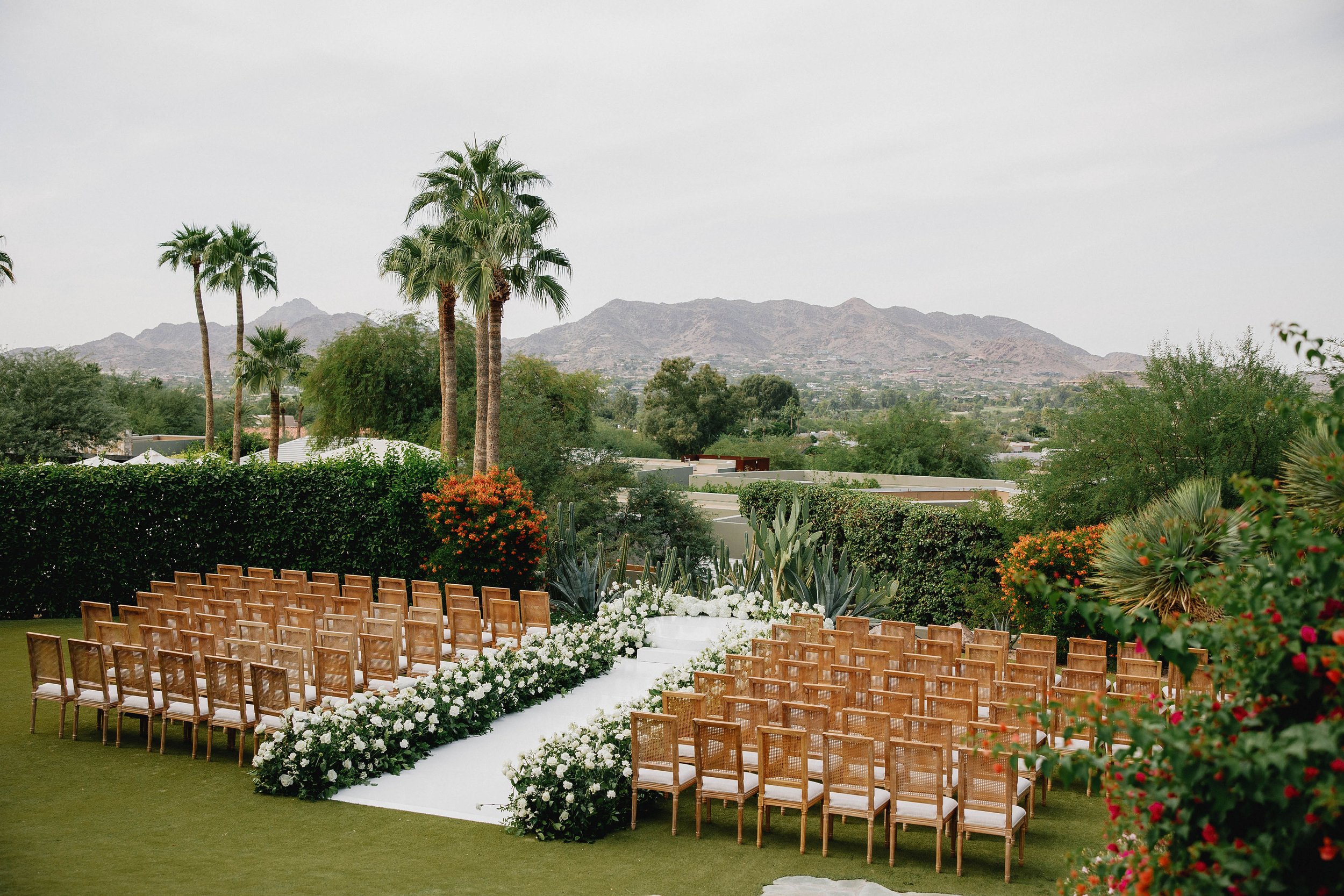 Destination wedding ceremony with white modern florals, wooden chairs, and lush greenery in an elegant garden setting
