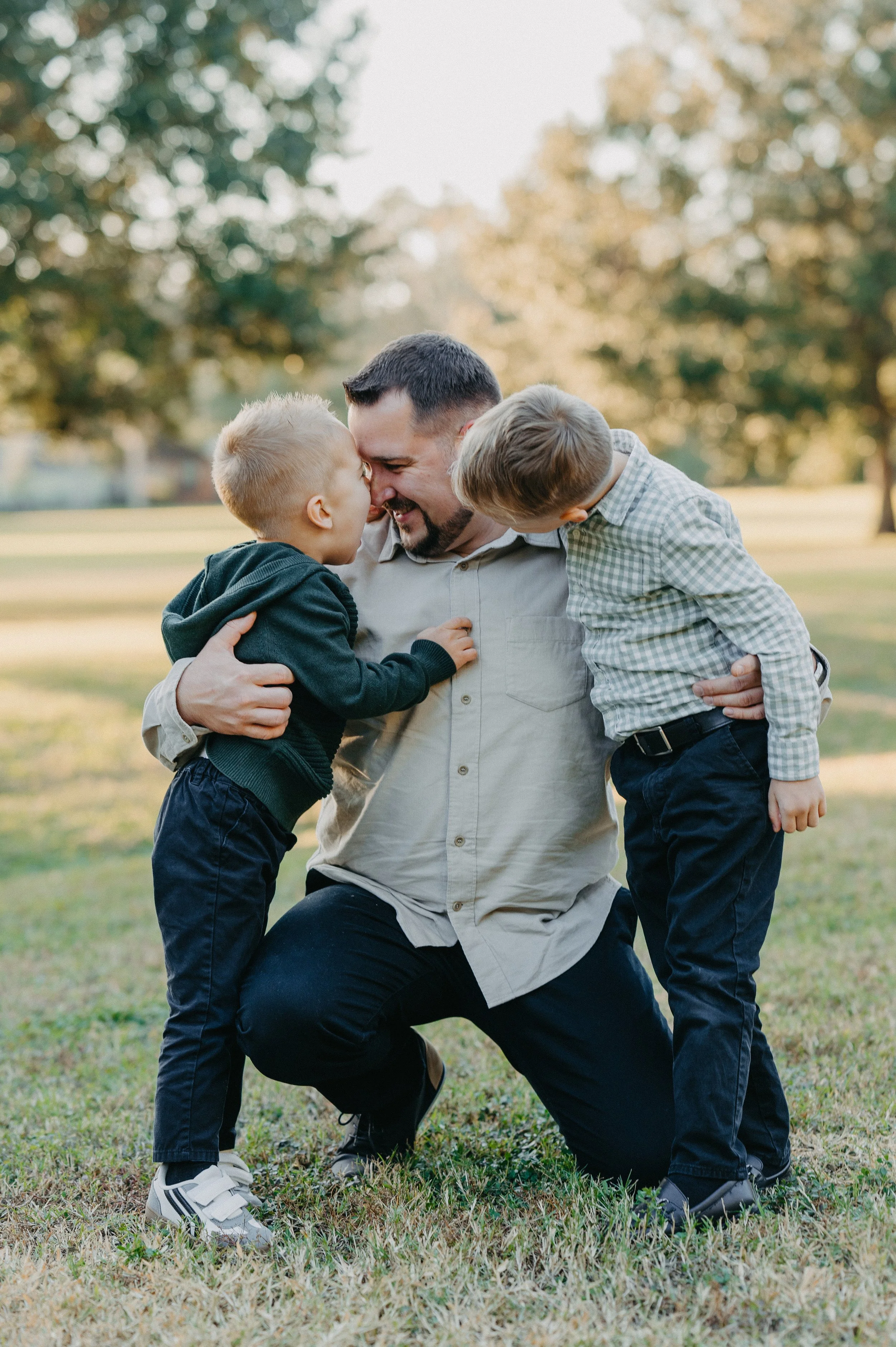 A man with two young boys outdoors, nose-to-nose, smiling, with trees in the background.