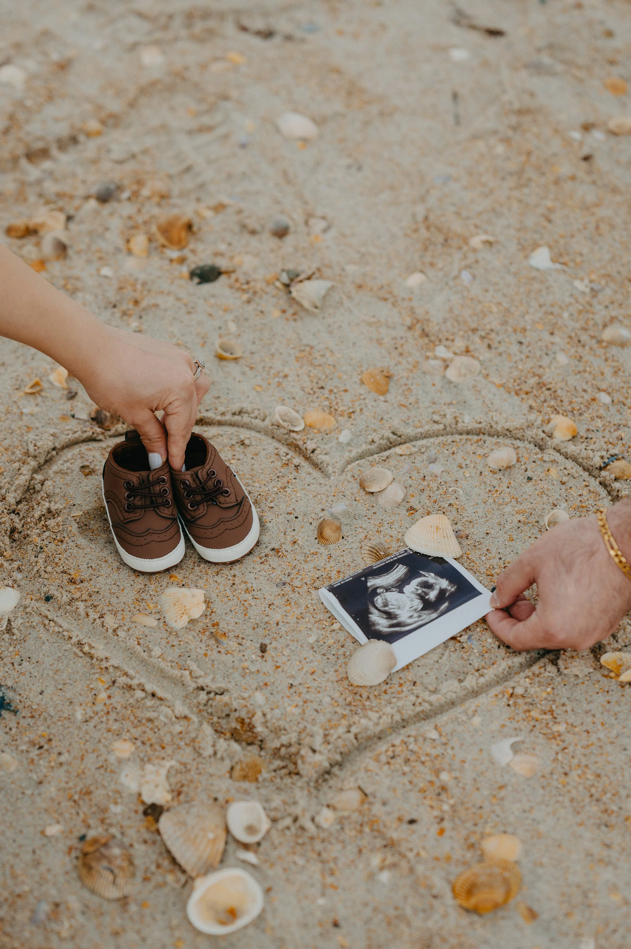 A baby's ultrasound picture and small brown shoes are placed in a heart shape drawn in the sand on the beach, surrounded by seashells.