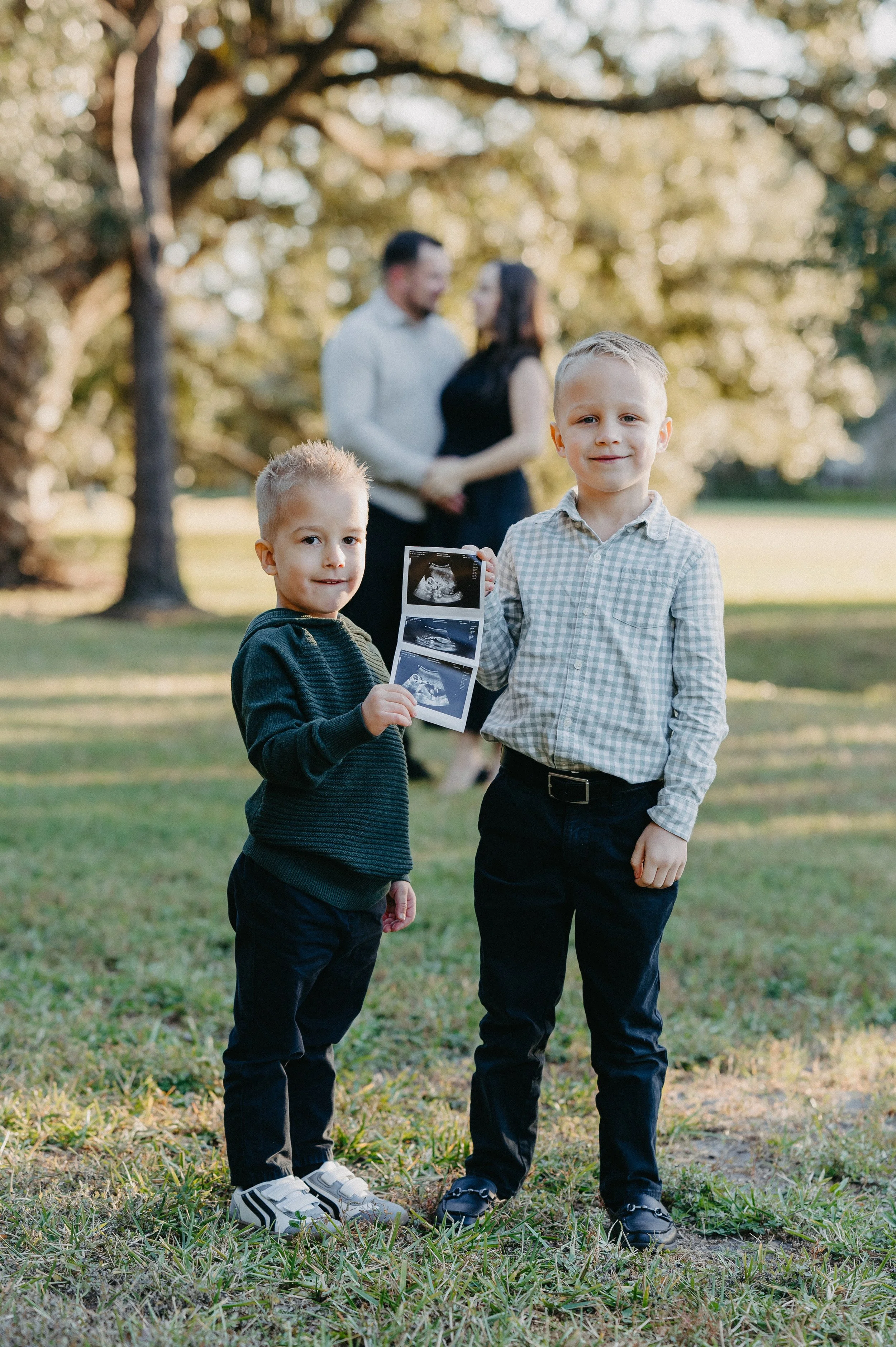 Two young boys standing outside in a park, holding ultrasound images, with a blurred couple embracing in the background.