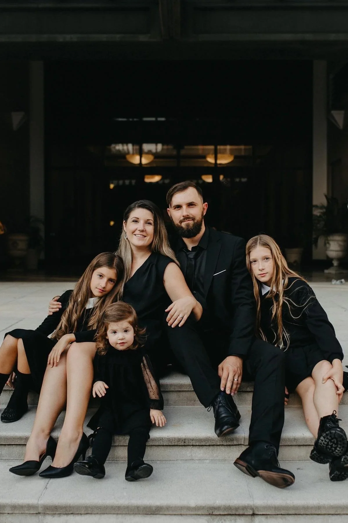 A family of six sitting on stairs outside a building at night, dressed in black formal attire, smiling at the camera.