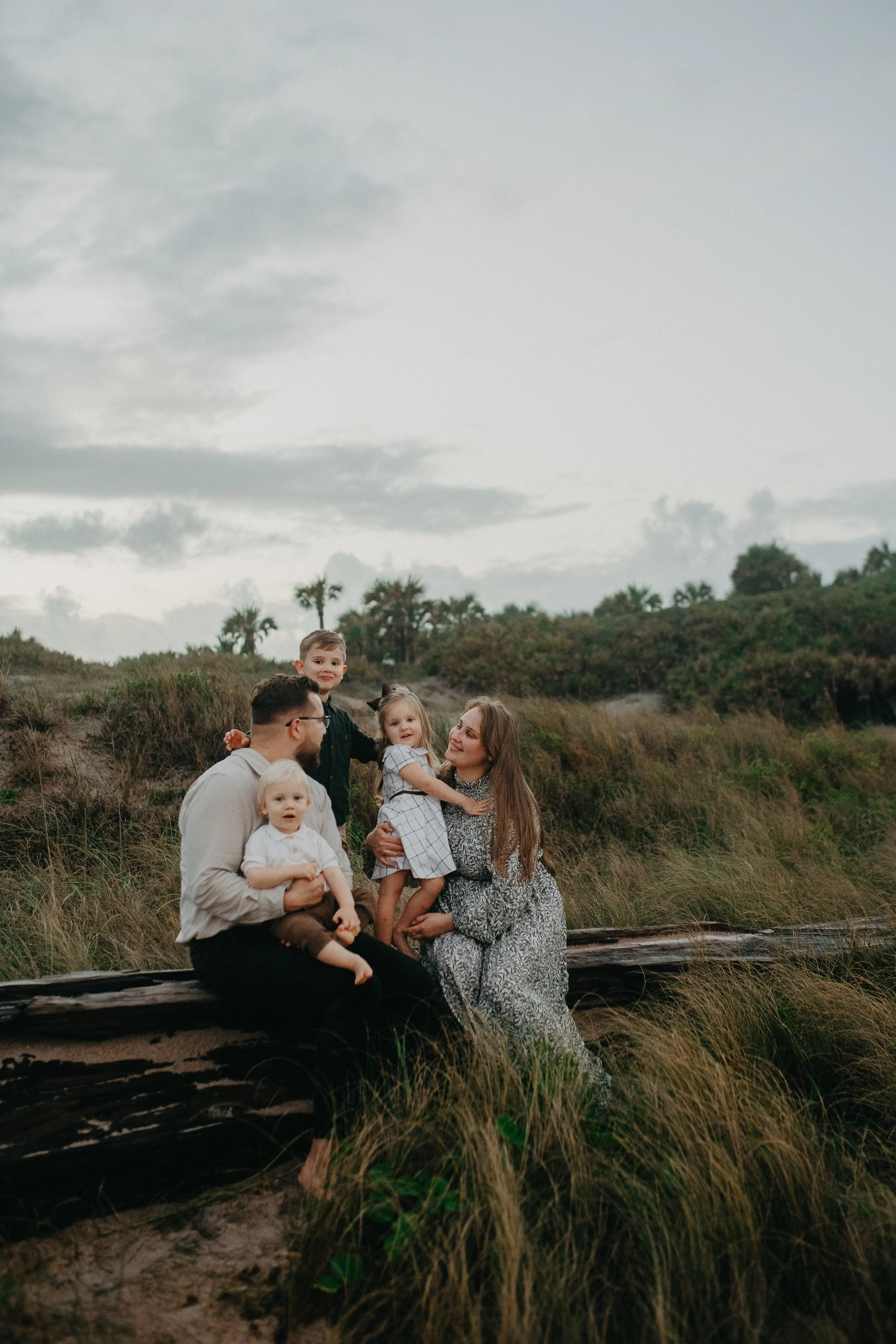 A family of five, including two parents and three young children, sitting on a log outdoors in a grassy, natural setting with a cloudy sky overhead.