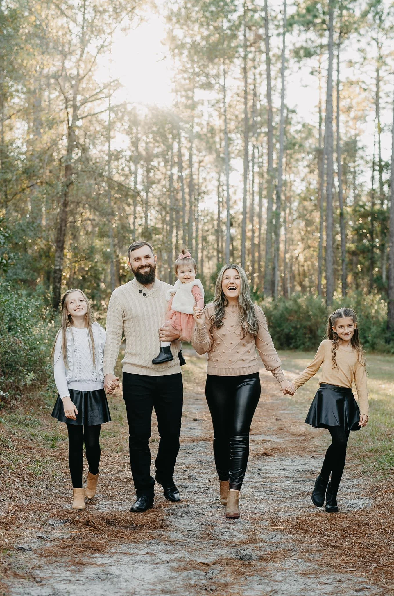 A family of five walking together on a wooded path in a forest, holding hands and smiling, with the sun shining through the trees.