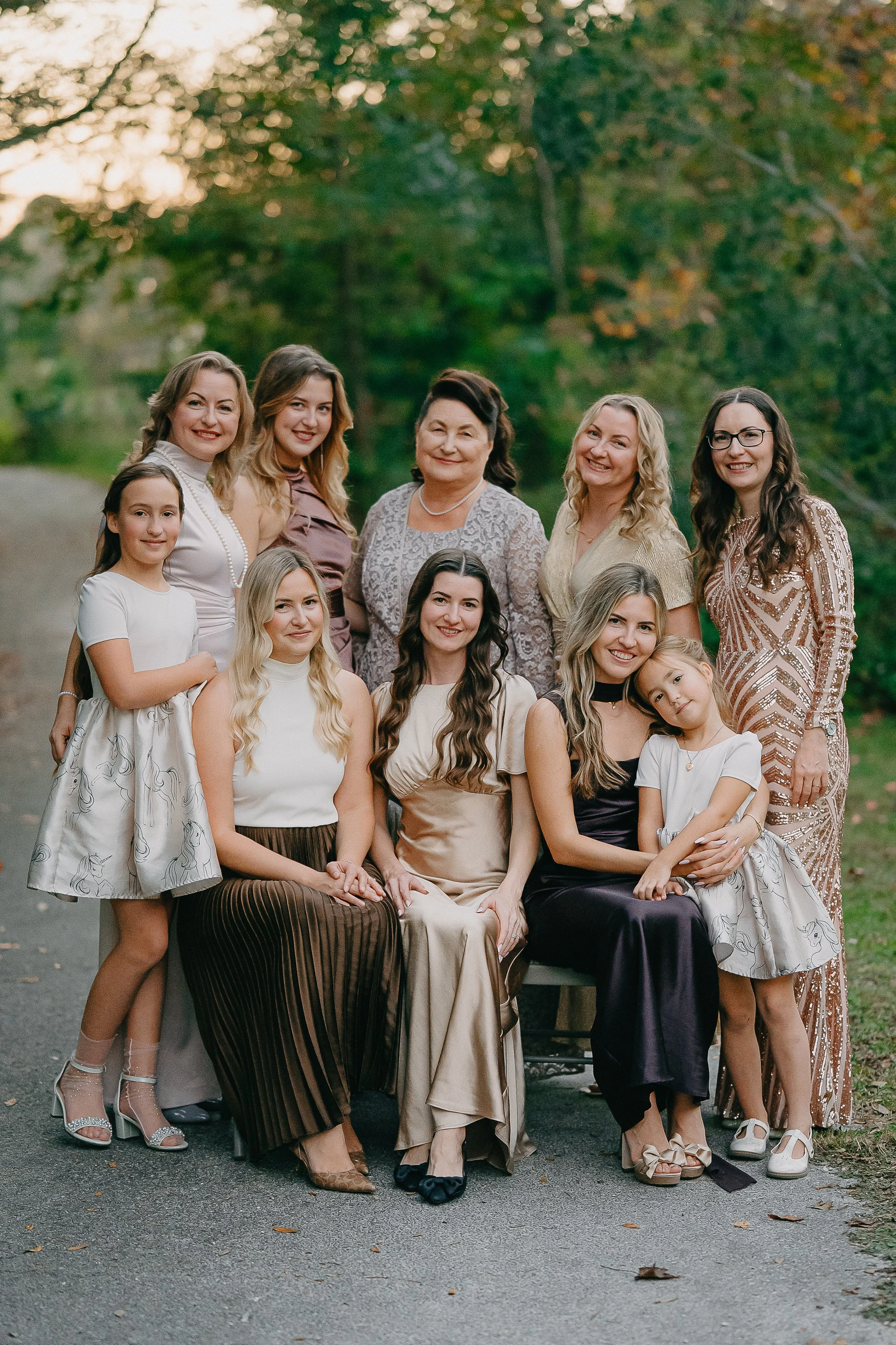 A group of twelve women and two young girls posing outdoors on a paved path with trees and greenery in the background, dressed in elegant attire for a family or social occasion.
