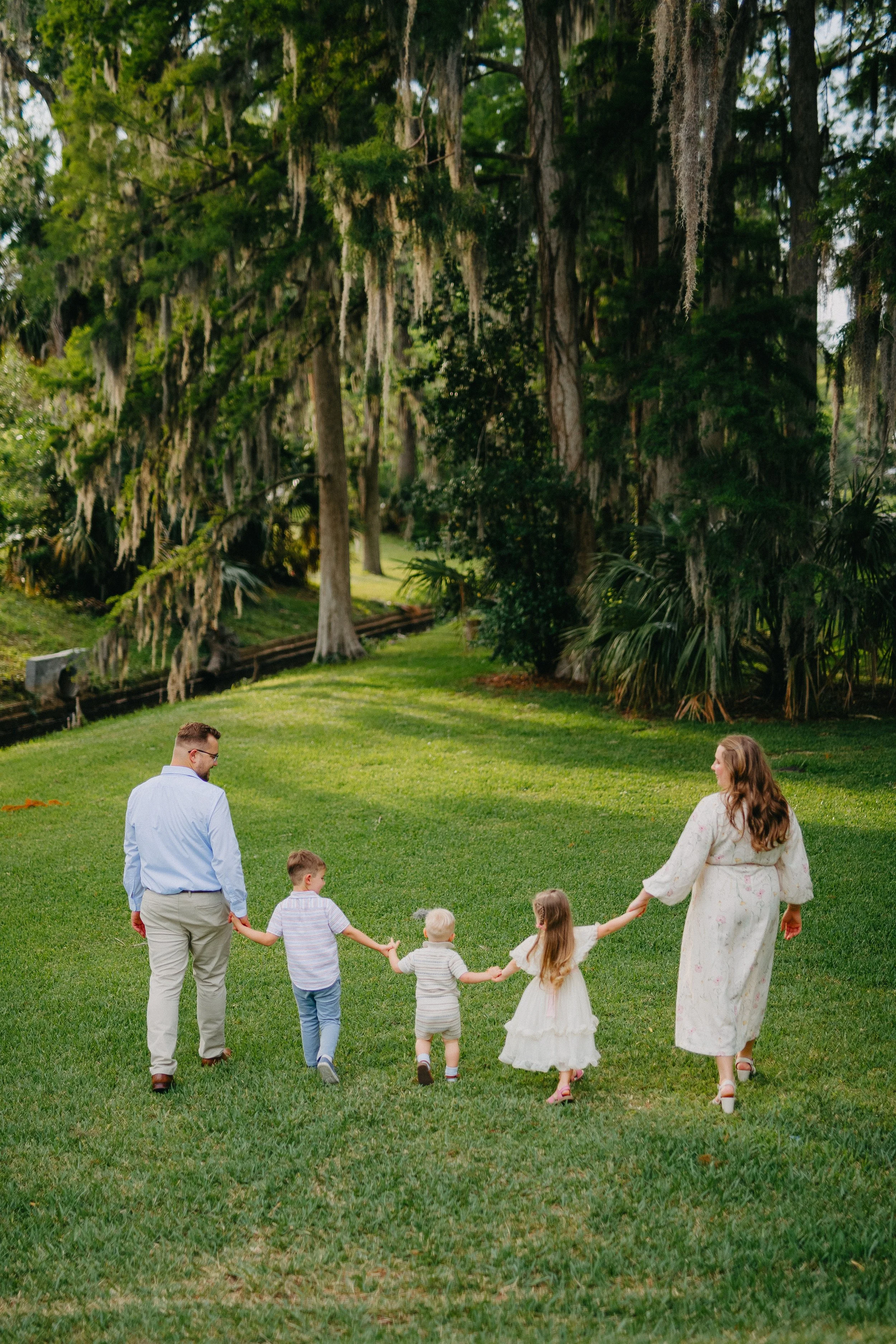 A family of six, including two adults and four children, walking hand in hand across a lush green lawn in a park surrounded by tall trees.