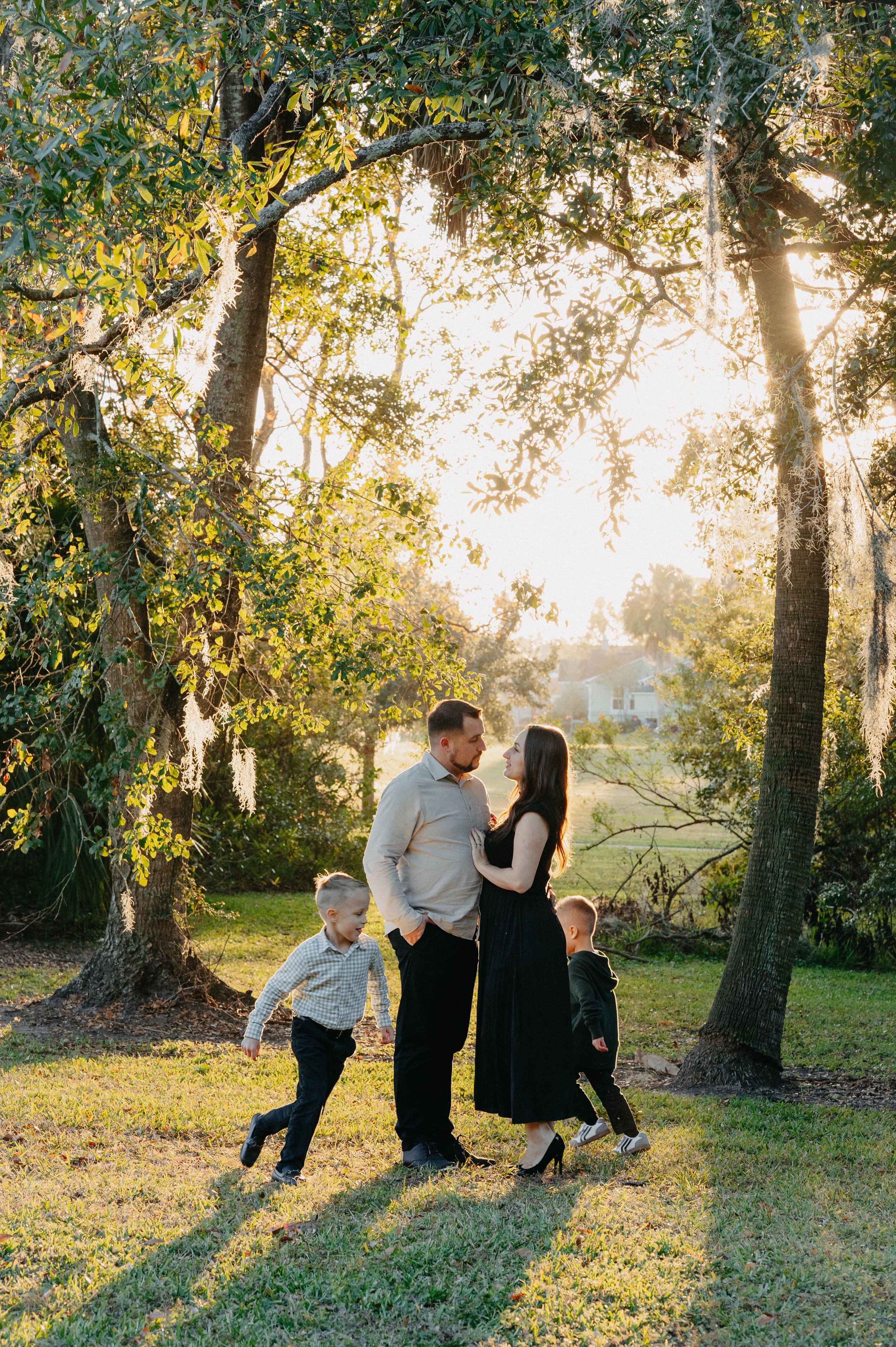 A family of four, including a man, a woman, and two young boys, stands together outdoors in a park during sunset, surrounded by trees and green grass.