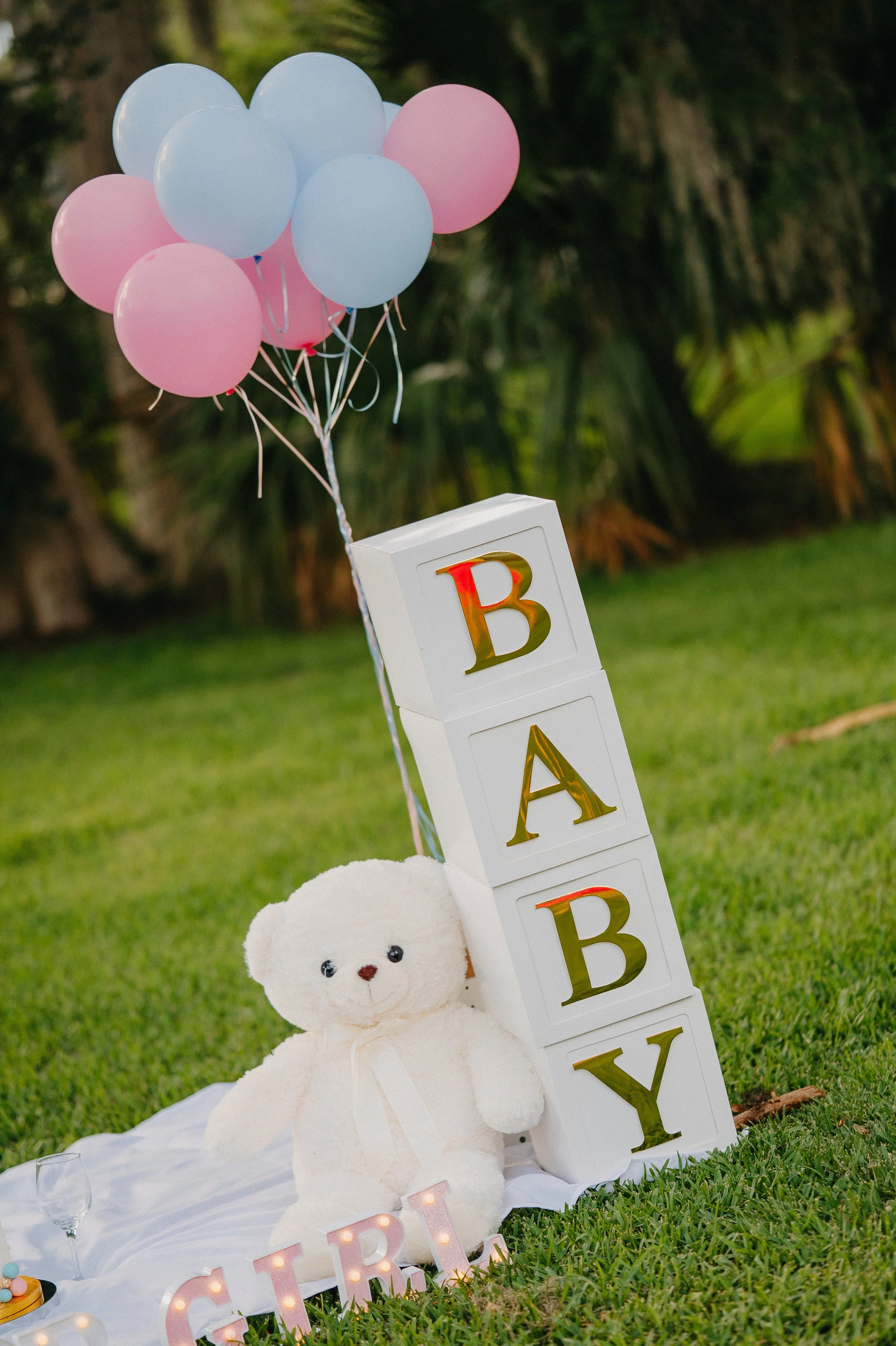 Decorative setup for a baby shower, including a teddy bear, pastel pink and blue balloons, and blocks spelling 'BABY' on grass.