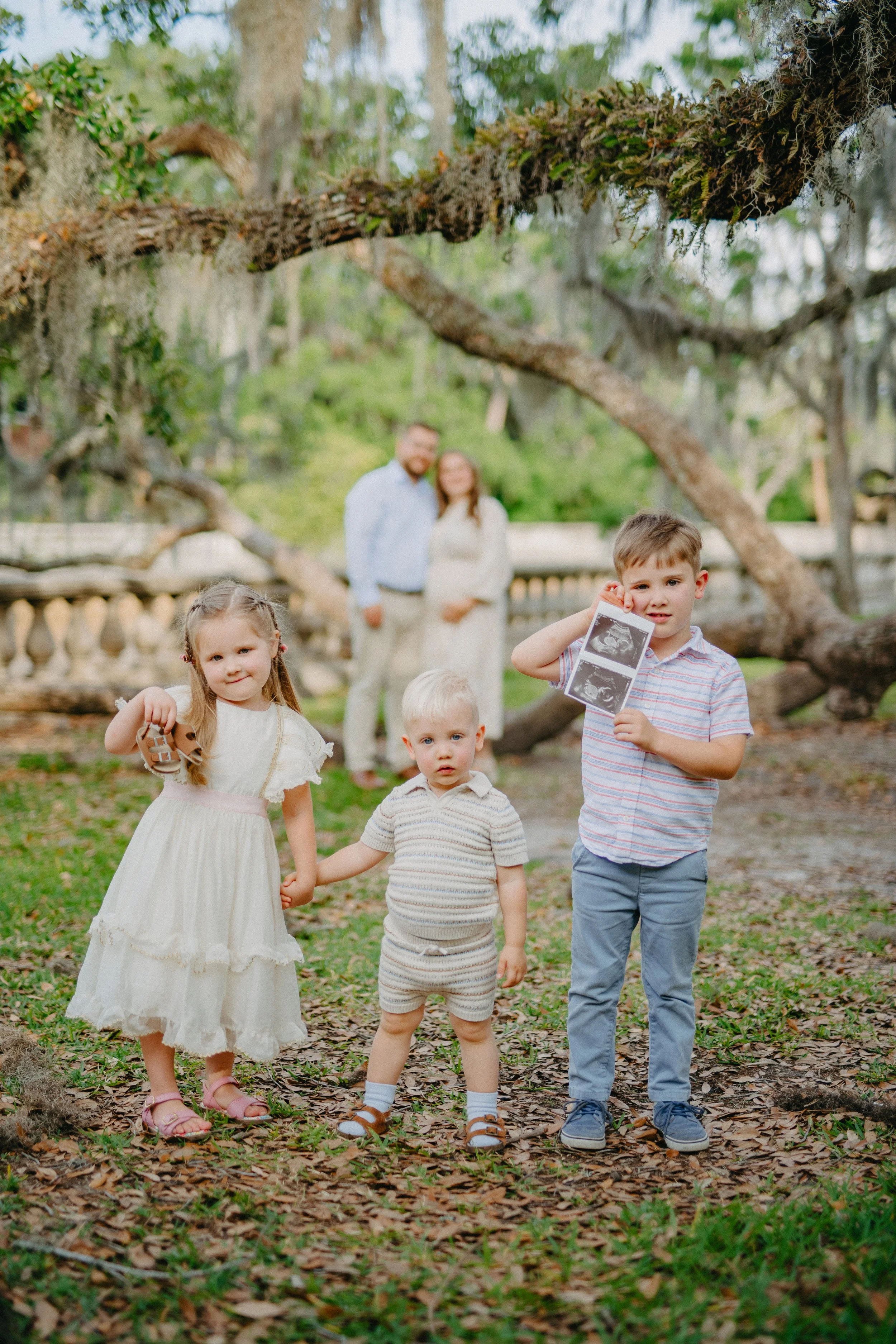 A family of five children standing outdoors in a grassy area with large tree branches overhead, with their parents blurred in the background; one young boy is holding ultrasound photos, another girl is holding a small stuffed animal, and two younger children are holding hands.