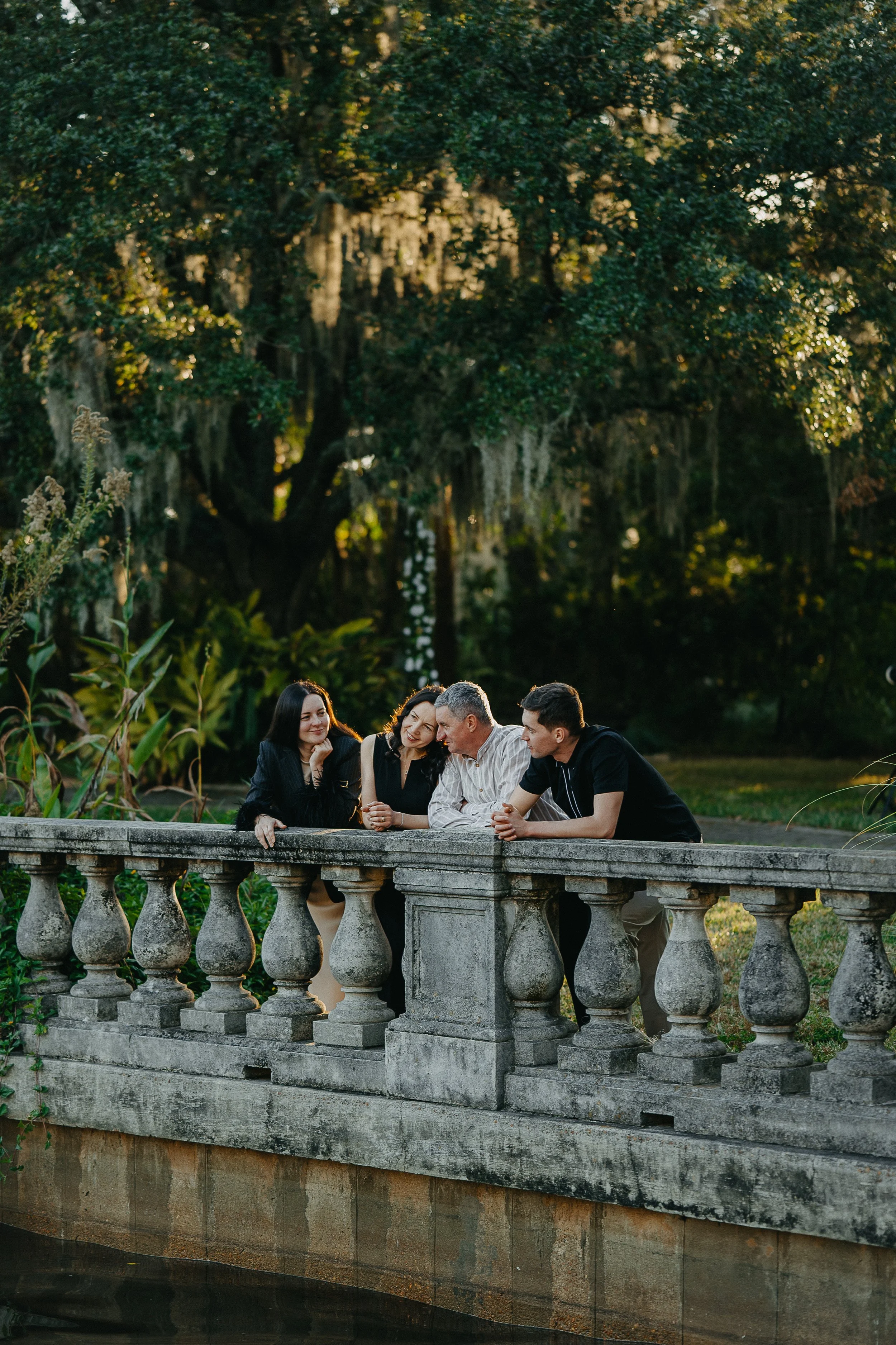 Four people standing on a stone bridge outdoors, engaged in conversation, with trees and greenery in the background.