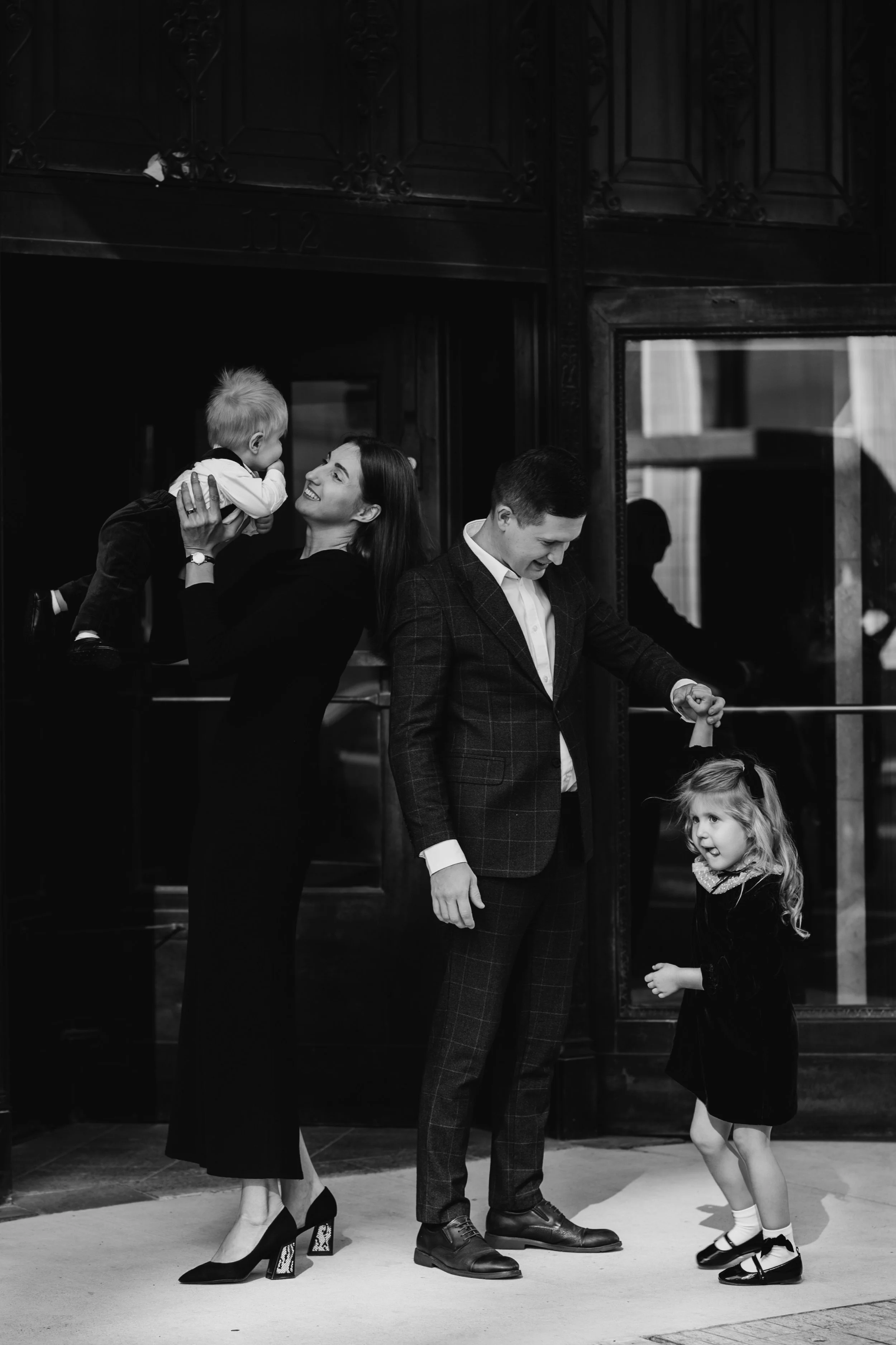 A family of four, including a woman, a man, a young girl, and a young boy, are inside a building near an entrance with dark wood and glass. The woman is holding a young boy up in the air, both smiling at each other. The man is smiling and looking down at the young girl, who is looking up at him, with a cheerful expression.