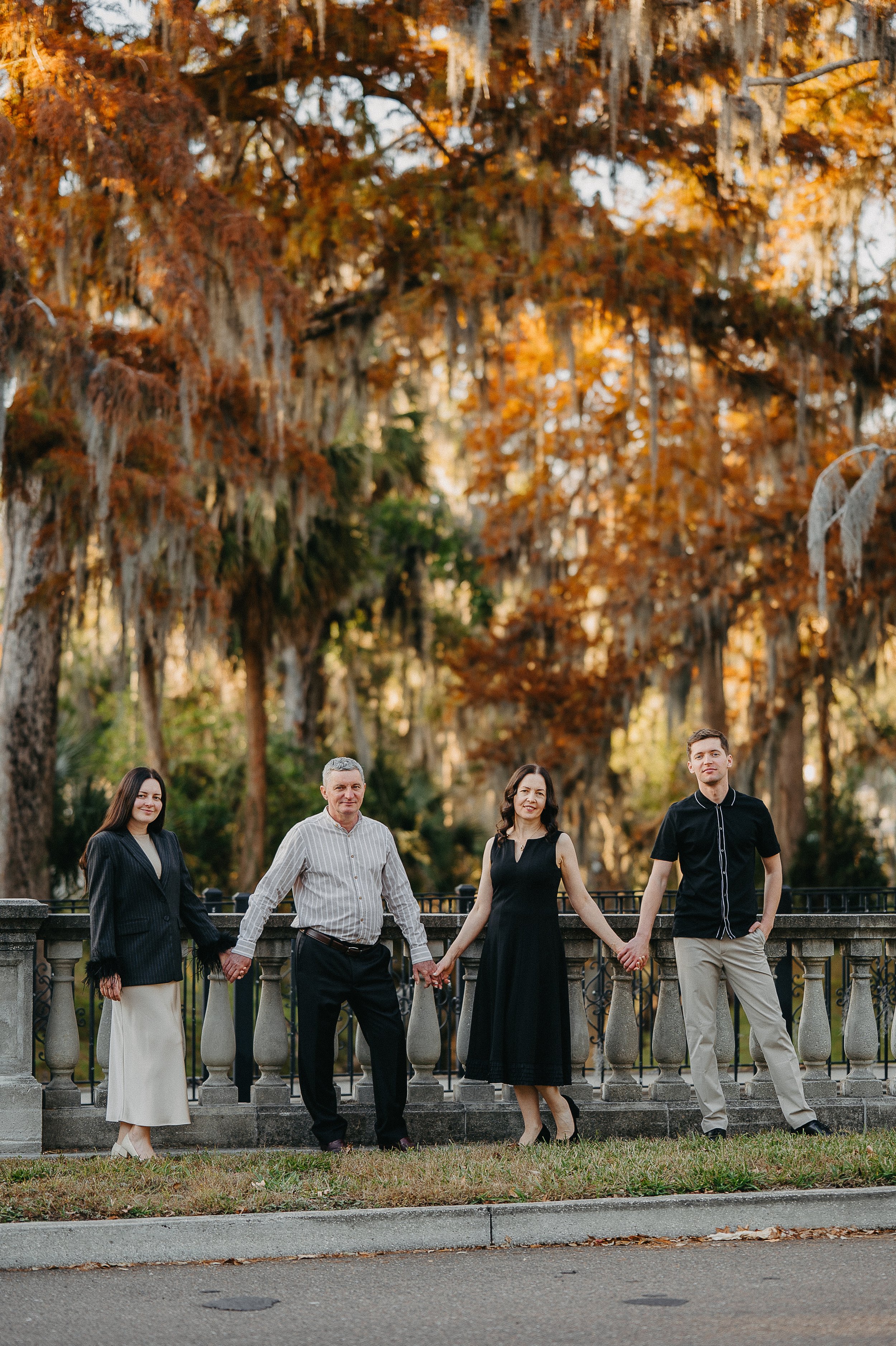 A family of four holding hands in a park with autumn trees in the background.