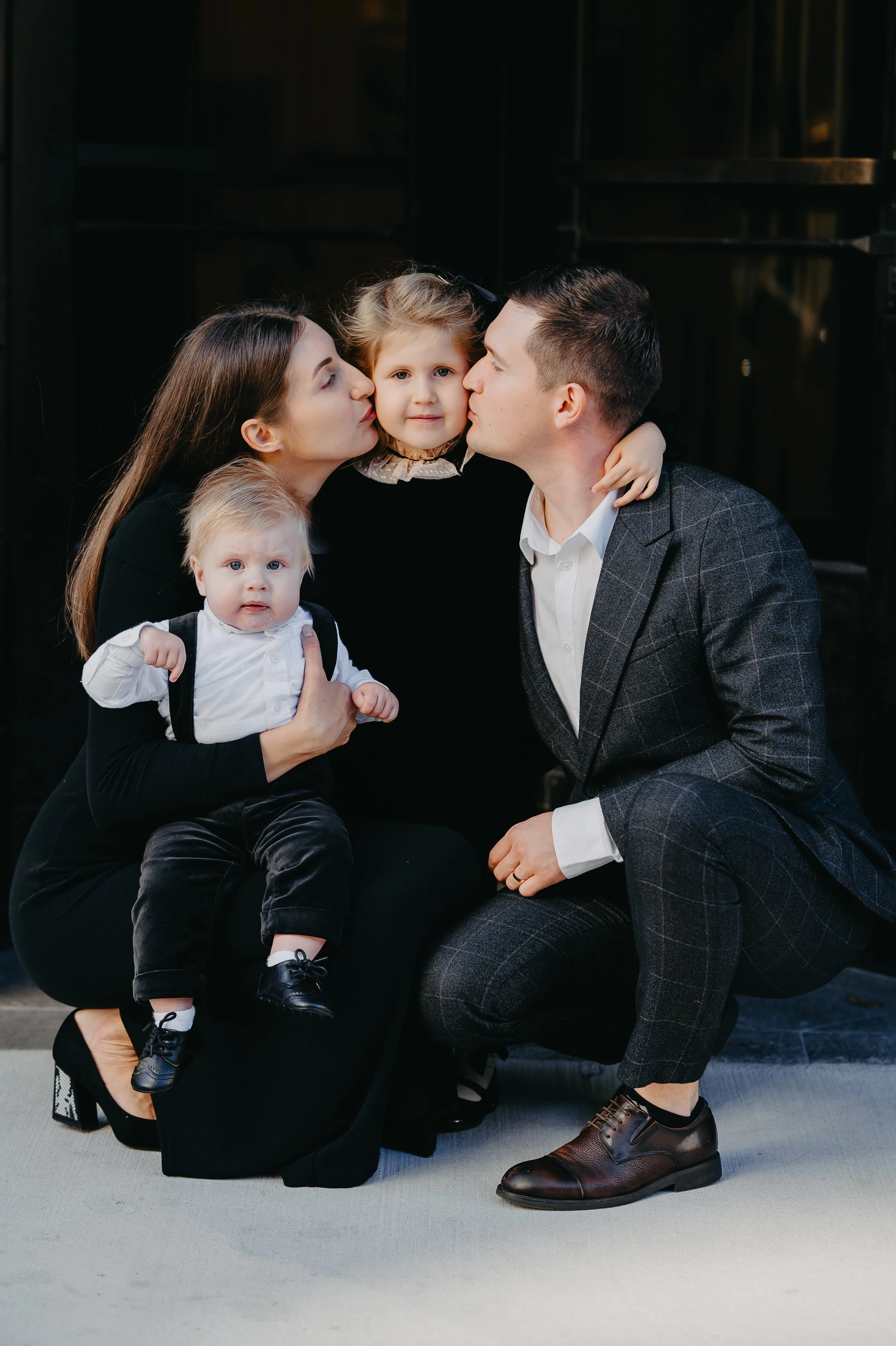 A family of four with a mother, father, and two young children, posed together. The mother holds a toddler, and the father kisses one of the children on the cheek, while the other child looks at camera. The background is dark and the family is dressed in formal attire.