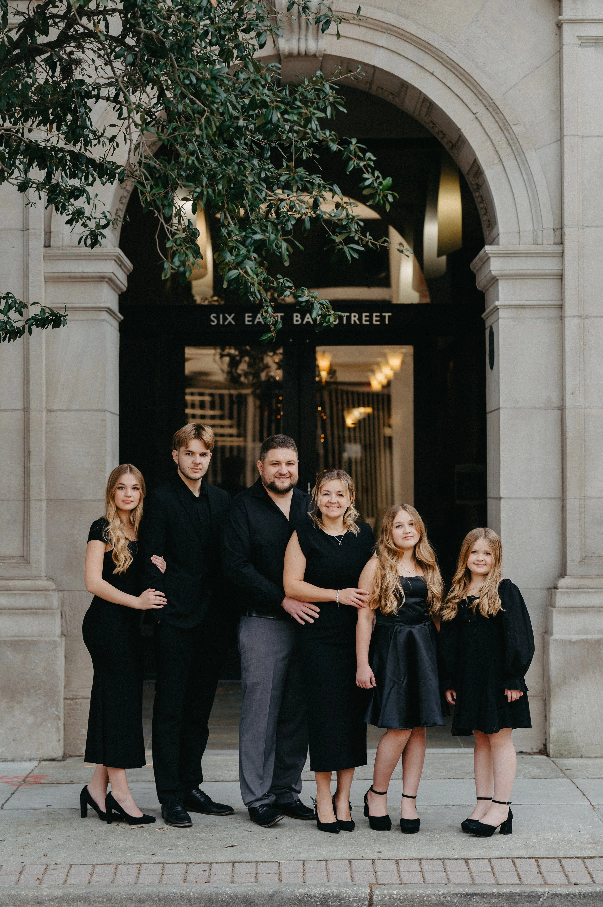 A family of six people standing in front of a building entrance, dressed in black. They are posing for a group photo with an archway behind them and the sign 'Six East Bae Street' above the entrance.