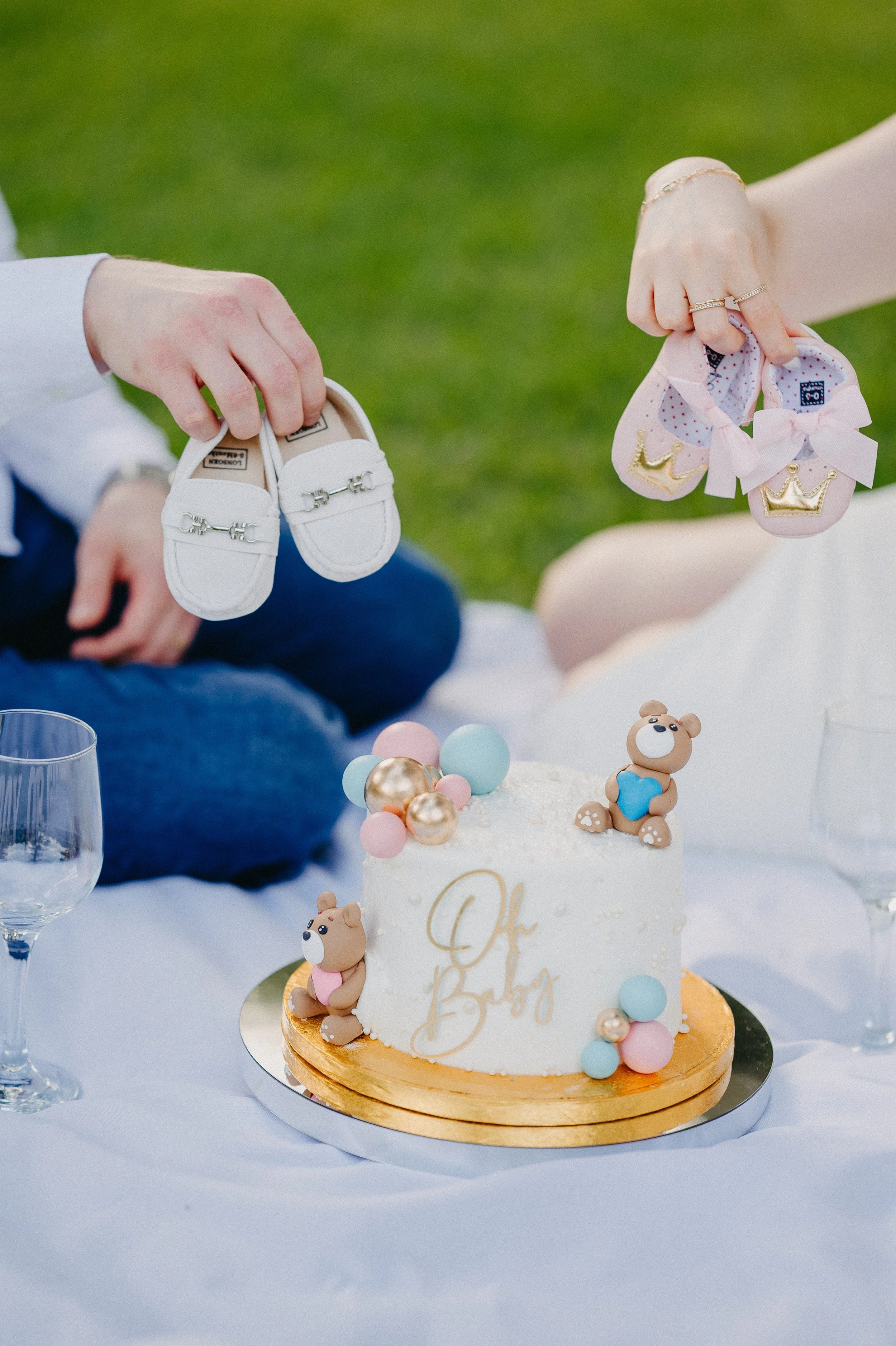 Baby shoes being held above a decorated birthday cake with teddy bear toppers, on a table with champagne glasses, during an outdoor celebration.