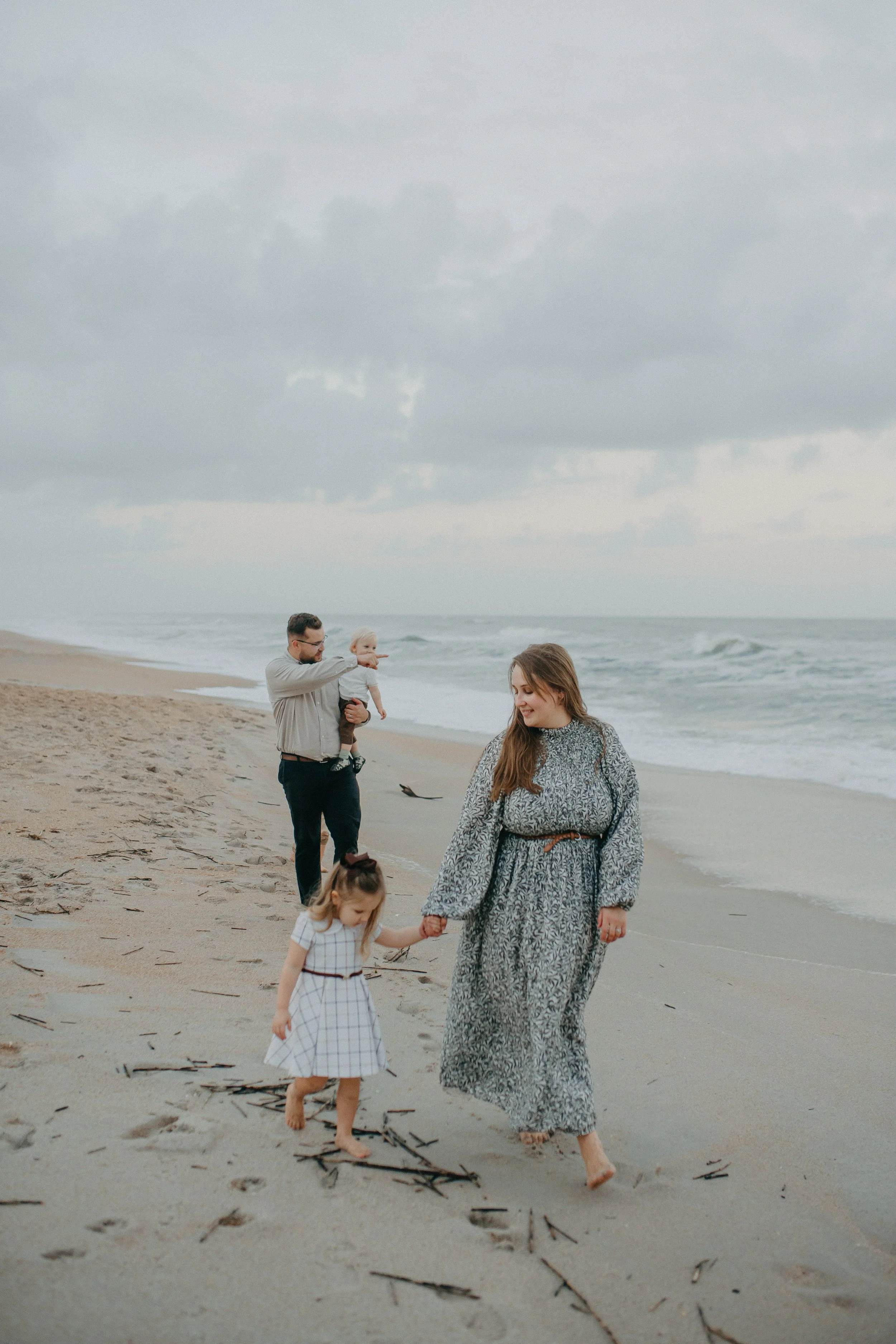 Family walking on a sandy beach, with a woman holding a young girl's hand and a man carrying a little boy, under a cloudy sky.