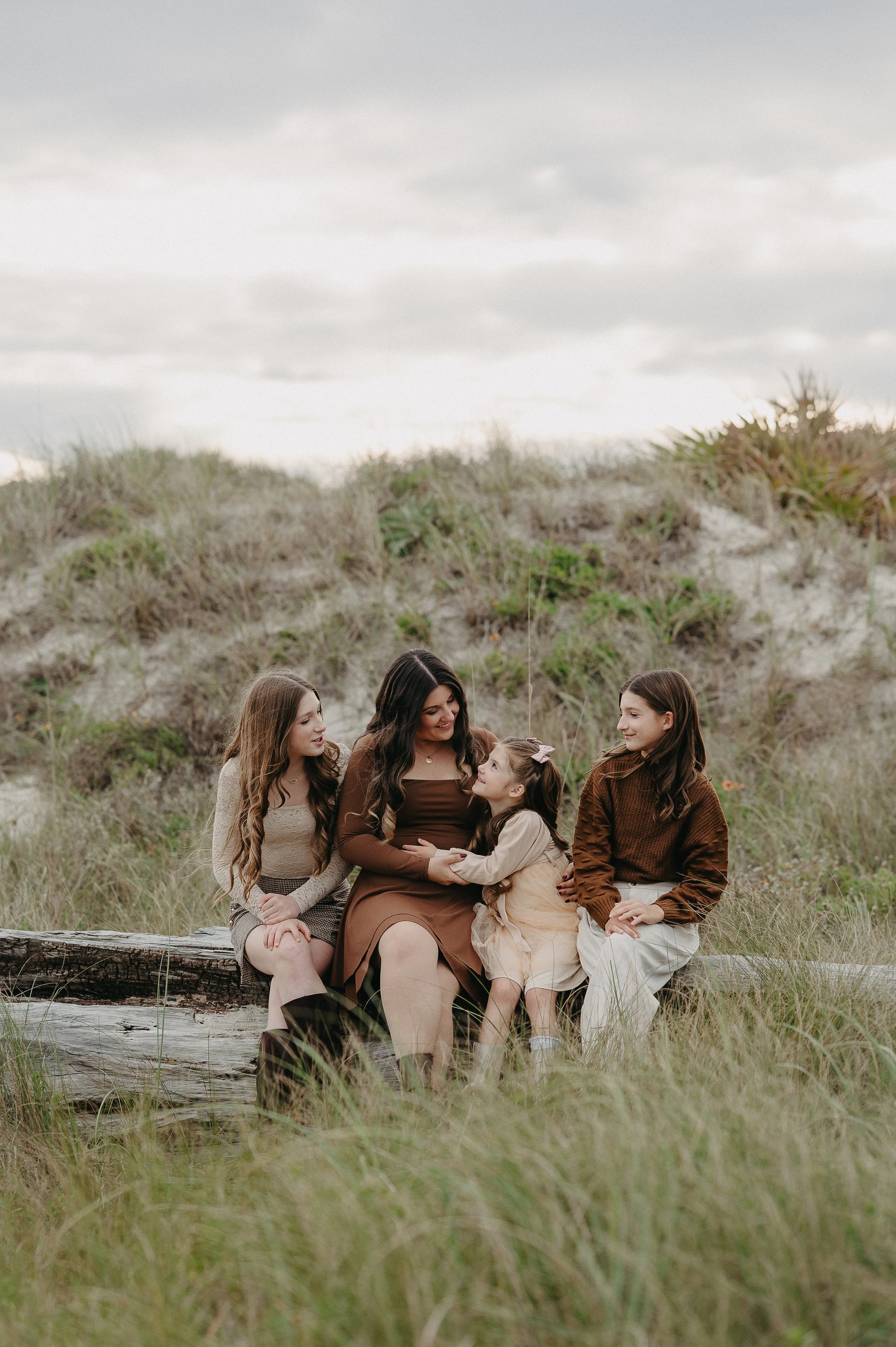 A woman with three young girls sitting on a log in a grassy outdoor area, engaging in a conversation during daytime with cloudy skies.