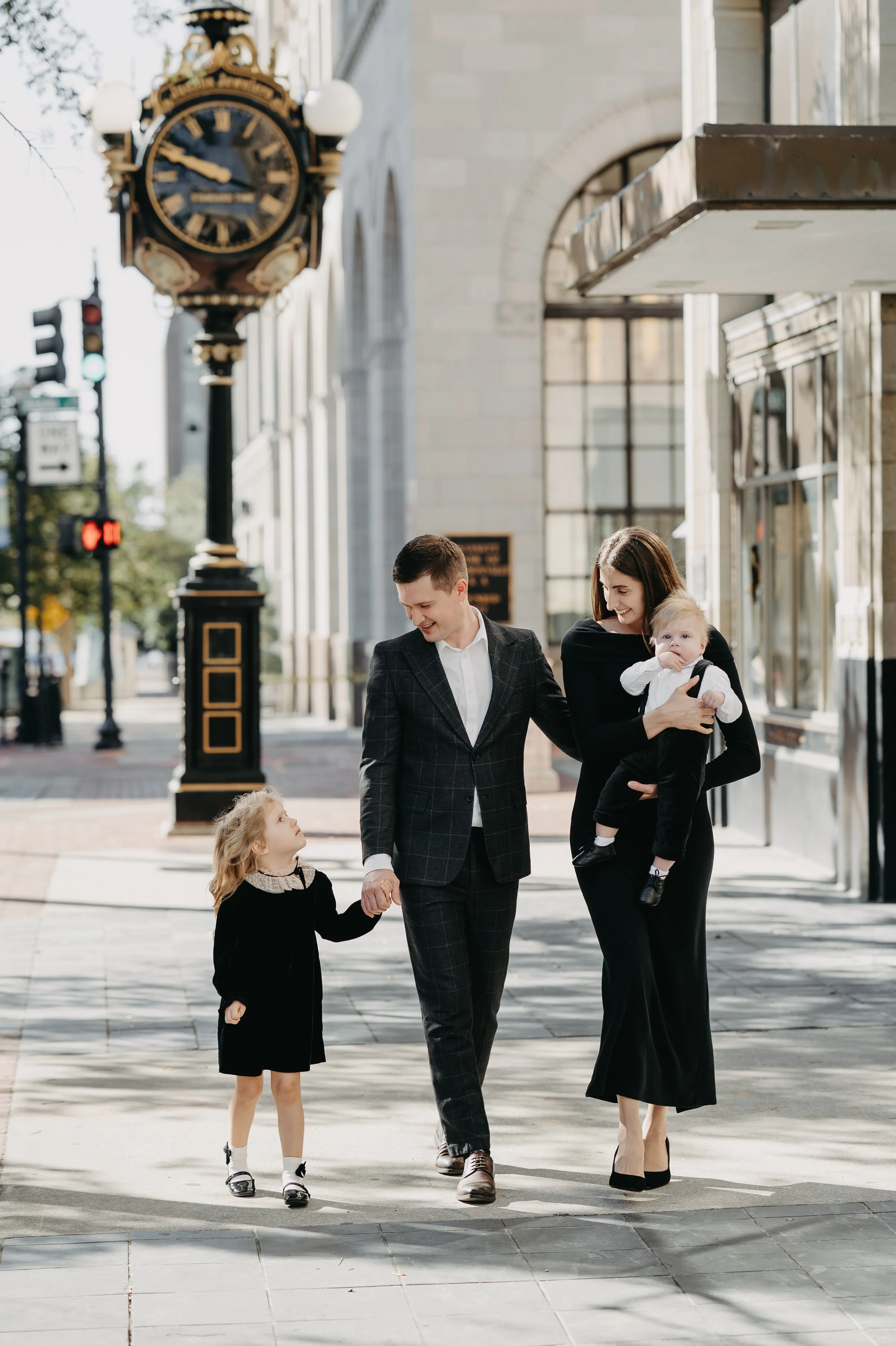 A family of four walking on a city sidewalk. The father is holding the hand of a young girl, and the mother is holding a young boy. The family is smiling and dressed in formal or semi-formal clothing.