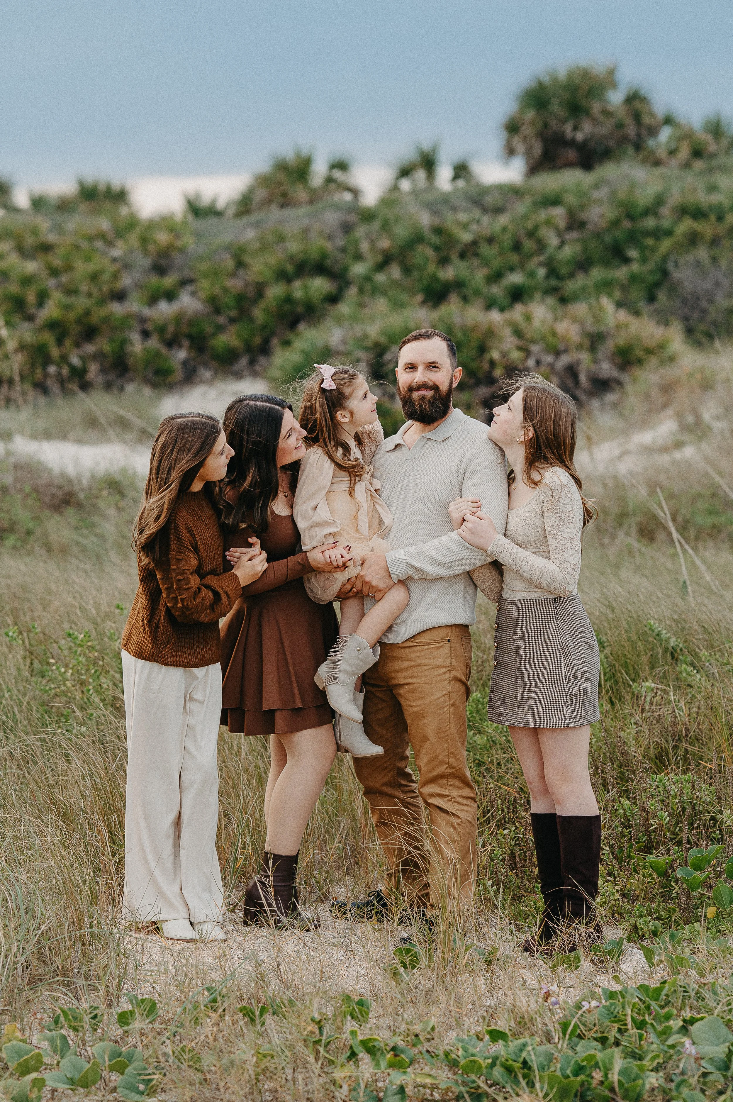 A man with a beard surrounded by four women and a girl in a grassy outdoor area with bushes and trees in the background.