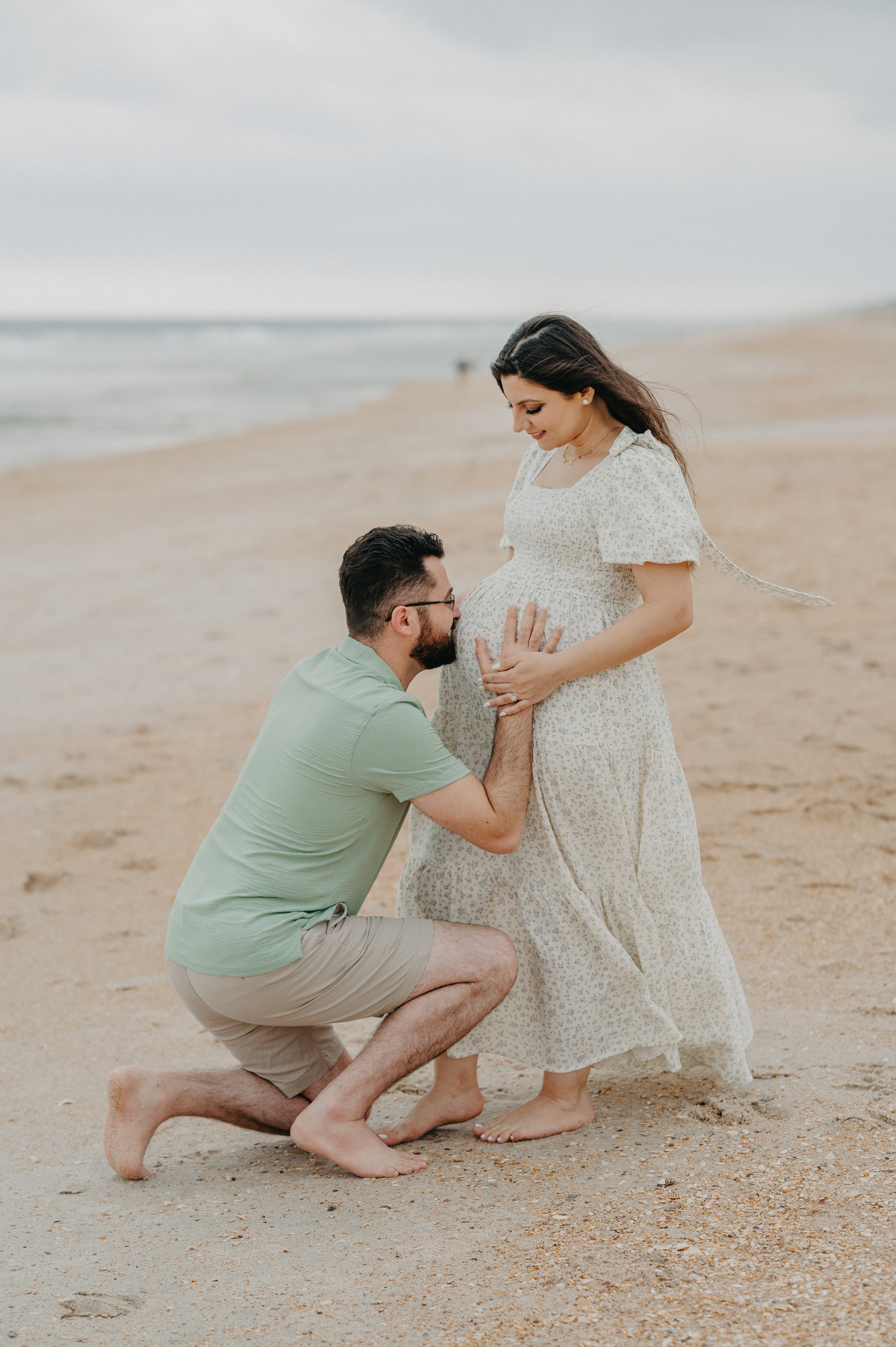A man kneels on the beach holding a pregnant woman's belly, who is standing in front of him, smiling and looking down at her belly. The woman wears a flowy, light-colored floral dress, and the man wears a light green shirt and beige shorts. The ocean is visible in the background.