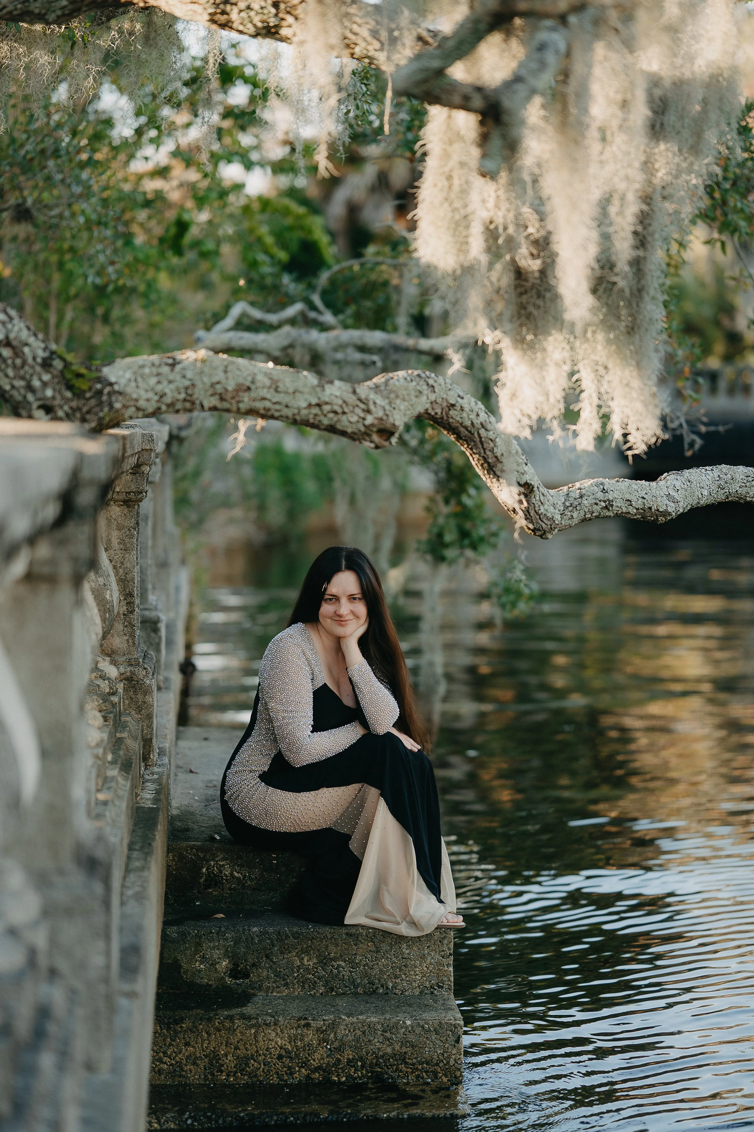 A woman with long dark hair sitting on stone steps beside a body of water, surrounded by trees with hanging moss, during late afternoon or early evening.