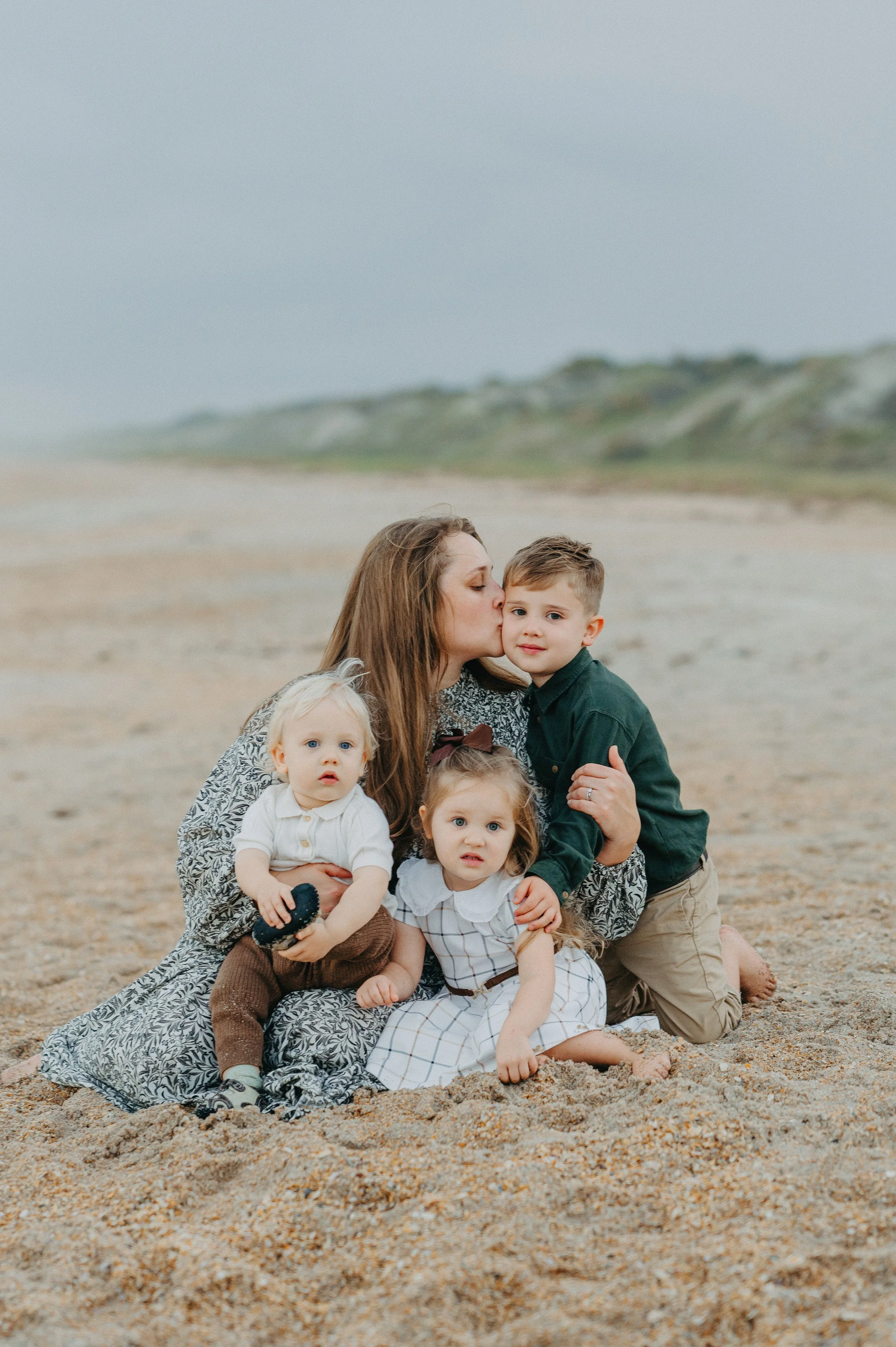 A woman and three young children on a beach, with the woman kissing a boy on the cheek, two girls sitting on sand, and a boy kneeling beside them.